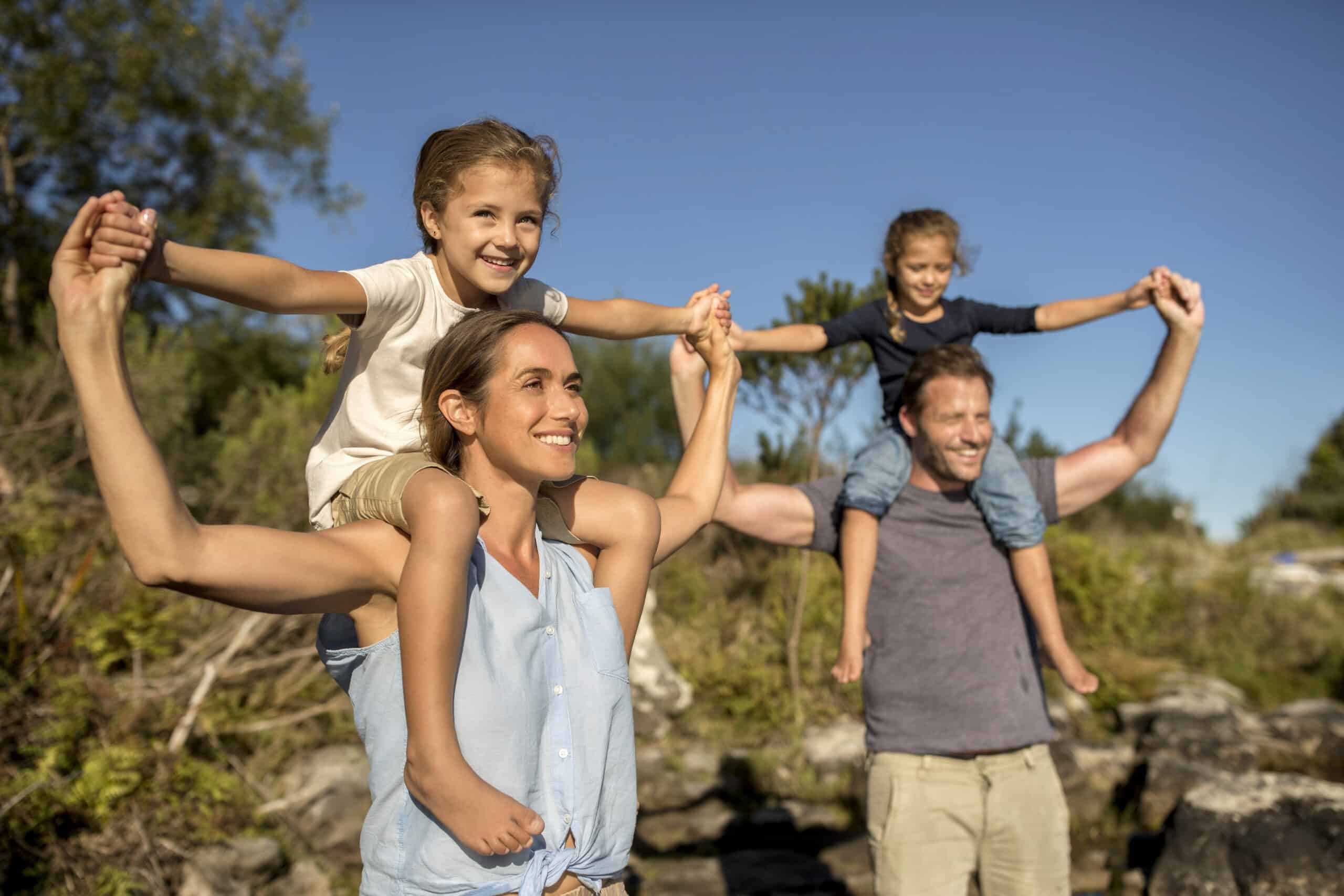 Eine fröhliche Familie im Freien mit blauem Himmel und Grün im Hintergrund. Die Eltern tragen ihre beiden kleinen Kinder auf den Schultern und strecken ihre Hände in die Luft. Die Familie wirkt glücklich und genießt gemeinsam einen lustigen Moment. © Fotografie Tomas Rodriguez