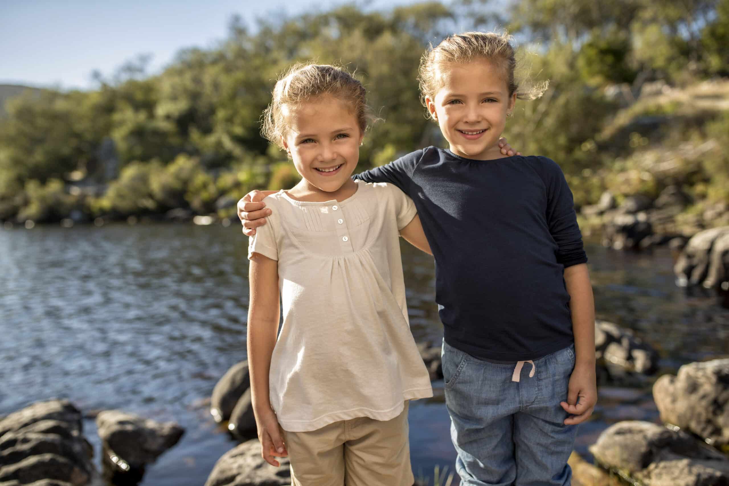 Zwei kleine Kinder stehen mit den Armen umeinander und lächeln in die Kamera. Sie sind an einem sonnigen Tag draußen an einem ruhigen See, umgeben von Felsen und Grün. Ein Kind trägt ein weißes Hemd, das andere ein blaues Hemd und eine blaue Hose. © Fotografie Tomas Rodriguez