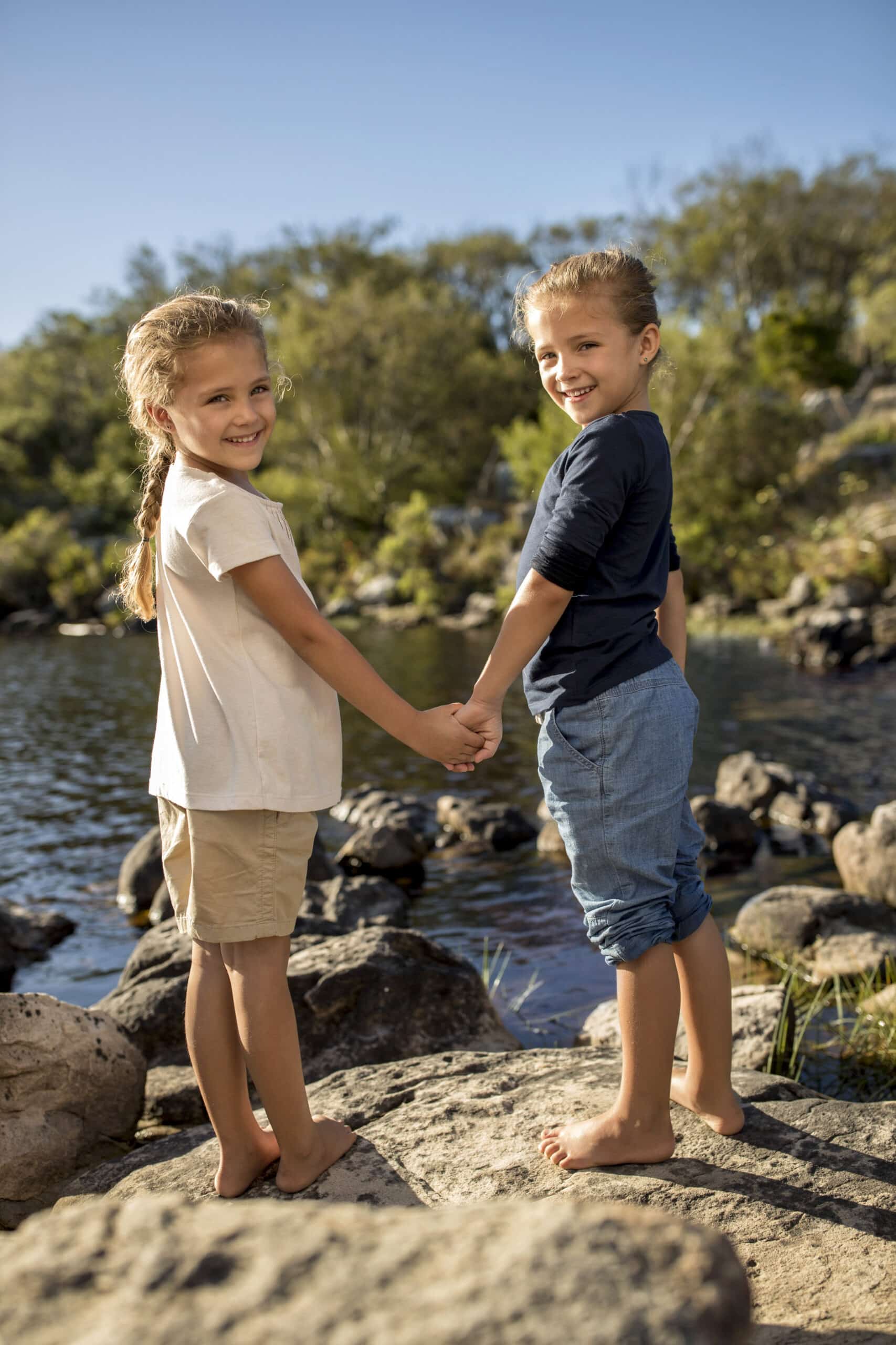 Zwei kleine Kinder, ein Junge und ein Mädchen, stehen barfuß auf Felsen an einem See, halten sich an den Händen und lächeln in die Kamera. Im Hintergrund sind Bäume und ein klarer blauer Himmel zu sehen, was einen sonnigen Tag in der Natur vermuten lässt. Beide Kinder wirken glücklich und entspannt. © Fotografie Tomas Rodriguez