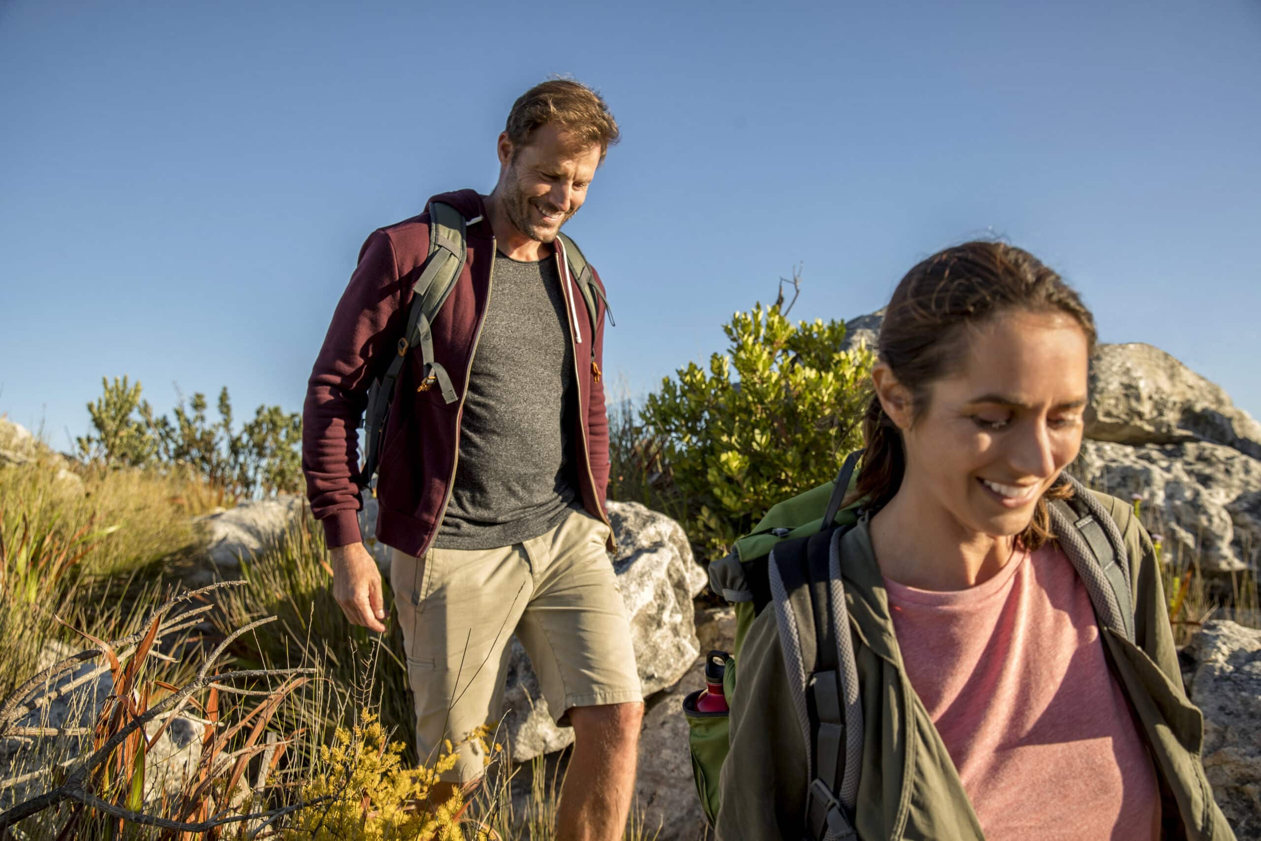 Ein Mann und eine Frau wandern durch eine felsige Landschaft mit Gras und Büschen. Der Mann, der eine kastanienbraune Jacke und beige Shorts trägt, lächelt und schaut nach unten. Die Frau, die ein rosa Hemd und eine grüne Jacke trägt, geht weiter und lächelt ebenfalls. Der Himmel ist klar und blau. © Fotografie Tomas Rodriguez