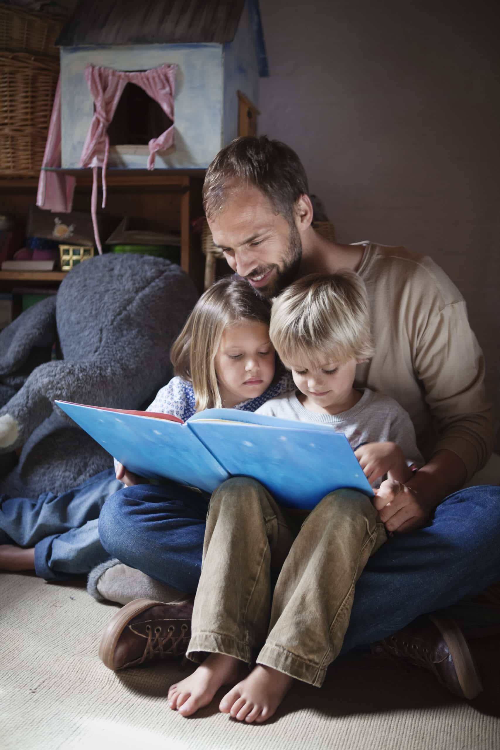 Ein bärtiger Mann sitzt mit einem blauen Buch auf dem Boden und liest zwei kleinen Kindern vor, die auf seinem Schoß sitzen. Sie befinden sich in einem gemütlichen Raum mit einem Spielzeughaus und einem großen Stofftier im Hintergrund. Die Kinder konzentrieren sich auf das Buch. © Fotografie Tomas Rodriguez