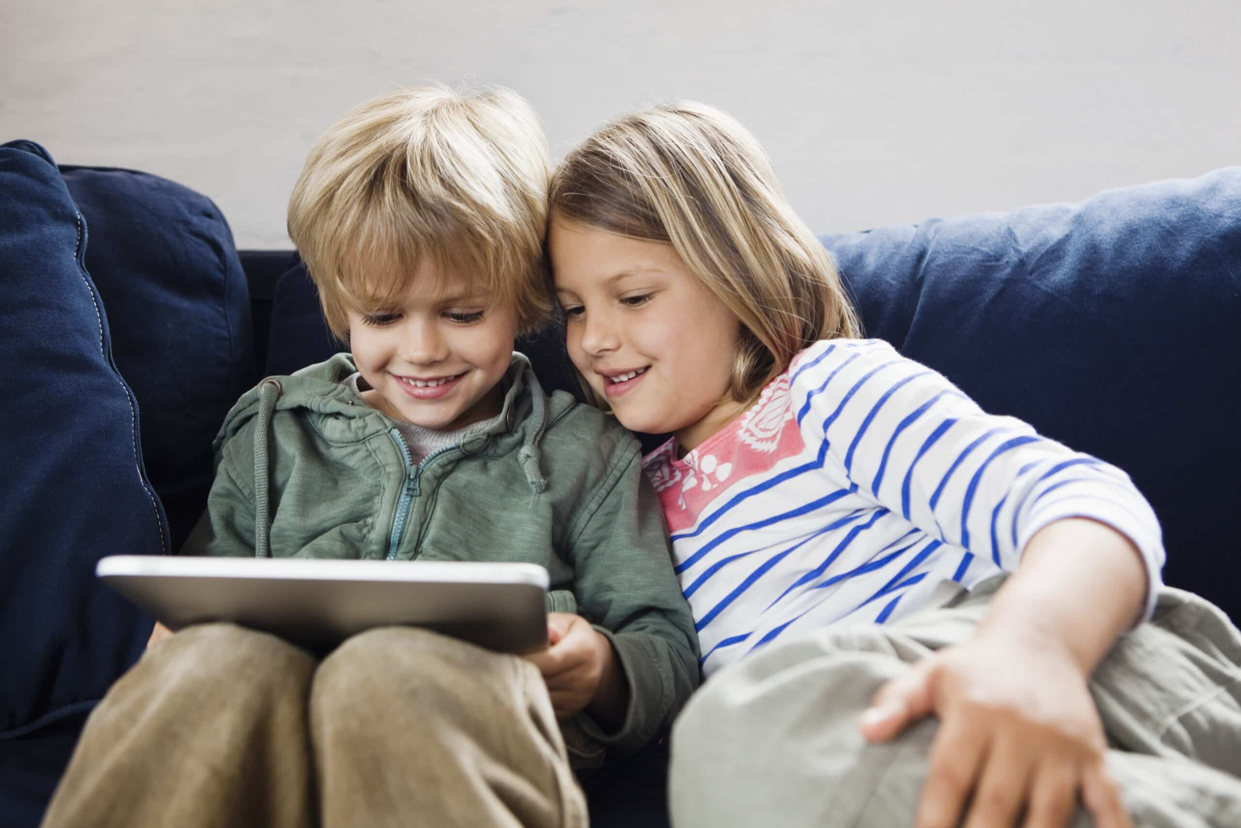 Zwei lächelnde Kinder sitzen eng beieinander auf einem dunkelblauen Sofa und schauen auf ein Tablet, das sie zusammen halten. Ein Kind trägt einen grünen Kapuzenpullover, das andere ein weiß-blau gestreiftes Shirt mit rosa Details. © Fotografie Tomas Rodriguez