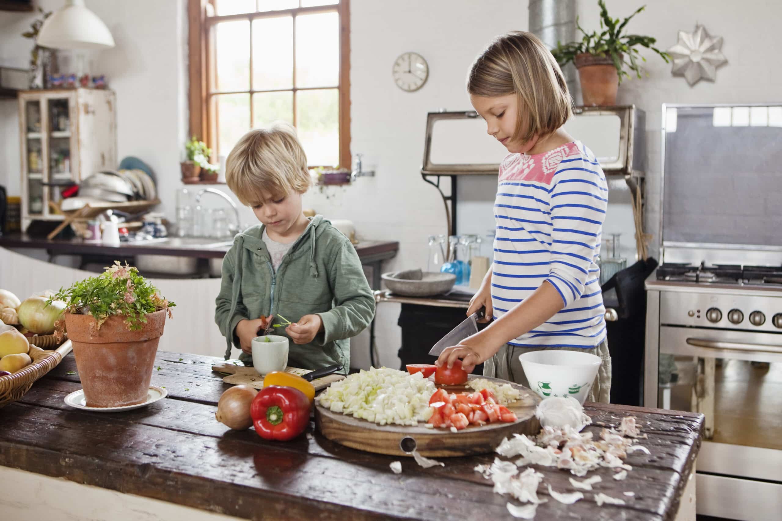 Zwei Kinder bereiten gemeinsam in einer Küche eine Mahlzeit zu. Der Junge links schält ein Gemüse, während das Mädchen rechts auf einer Holztheke Tomaten schneidet. Um sie herum liegen verschiedene Gemüsesorten und Utensilien verstreut, im Hintergrund sind Küchenutensilien zu sehen. © Fotografie Tomas Rodriguez