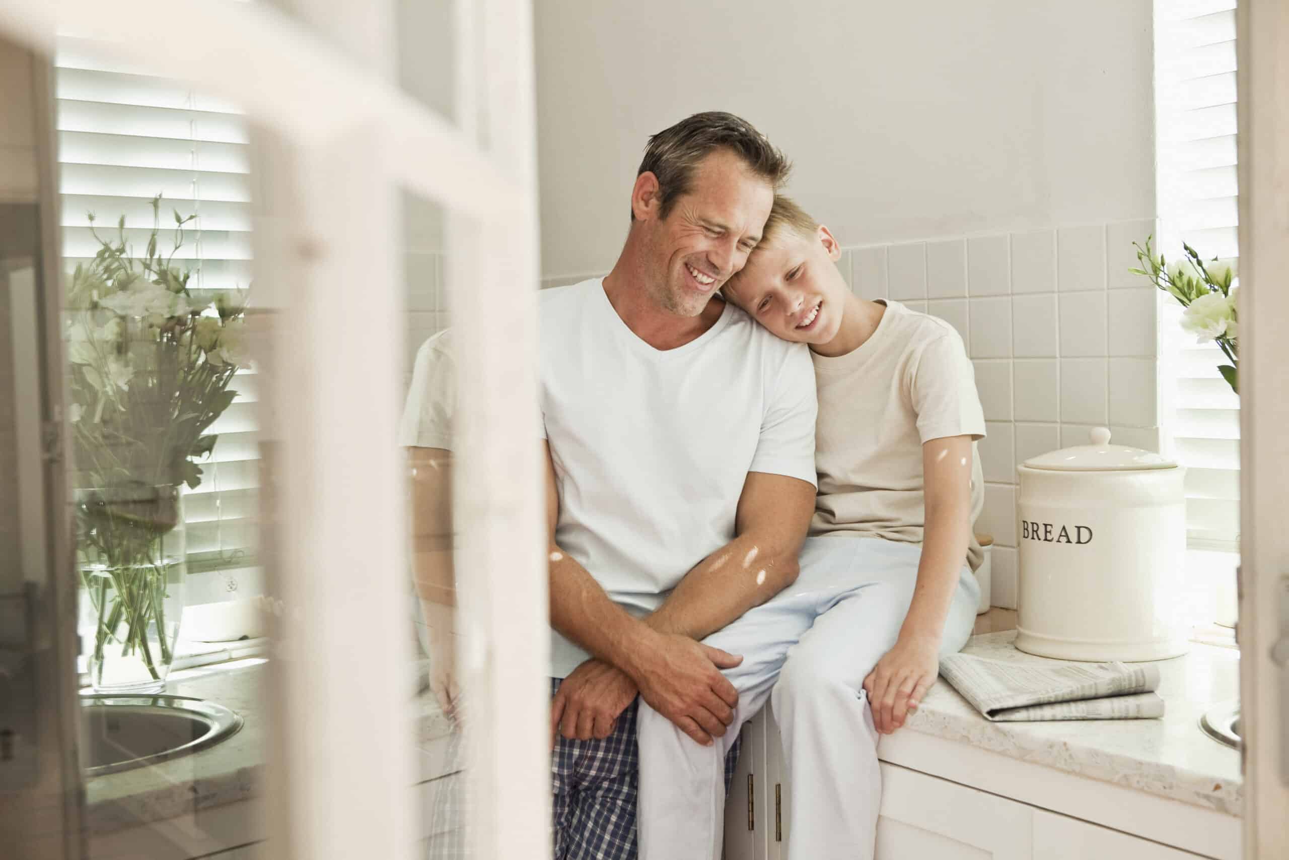 Ein Mann und ein Junge sitzen auf einer Küchentheke, beide lächeln und lehnen ihre Köpfe zusammen. Der Mann hat seine Arme um den Jungen gelegt. Sie tragen legere weiße Hemden. Die Küche hat eine ruhige Atmosphäre mit weißen Fliesen, einem Brotbehälter und Blumen im Hintergrund. © Fotografie Tomas Rodriguez