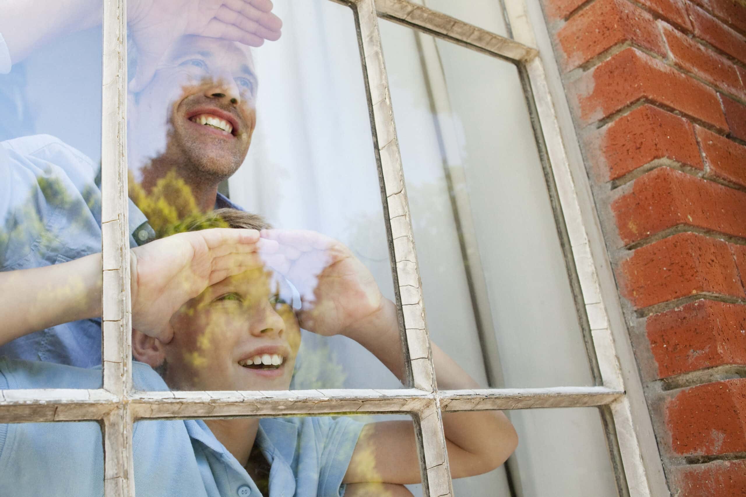 Ein Mann und ein kleiner Junge schauen aus dem Fenster und bedecken ihre Augen mit den Händen. Sie lächeln und scheinen die Aussicht zu genießen. Der Fensterrahmen ist alt und verwittert, und auf dem Glas spiegeln sich Sonnenlicht und Grün. © Fotografie Tomas Rodriguez