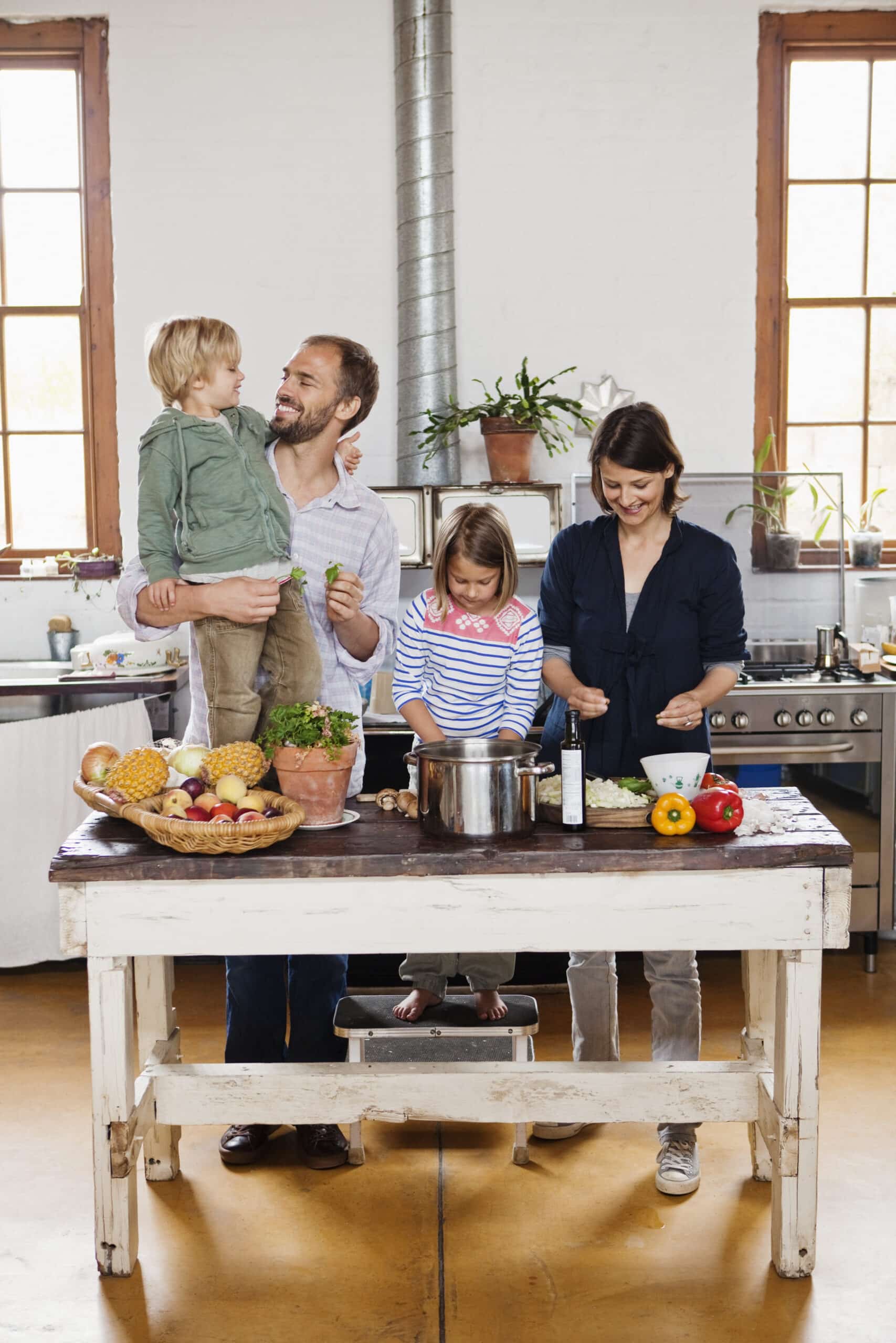Eine vierköpfige Familie steht in einer Küche und bereitet gemeinsam Essen zu. Ein Mann, der einen lächelnden jungen blonden Jungen im Arm hält, steht neben einer Frau, die an einer Theke Gemüse schneidet. Ein jüngeres Mädchen steht ebenfalls an der Theke und konzentriert sich auf einen Topf. Die Küche hat Pflanzen und große Fenster. © Fotografie Tomas Rodriguez