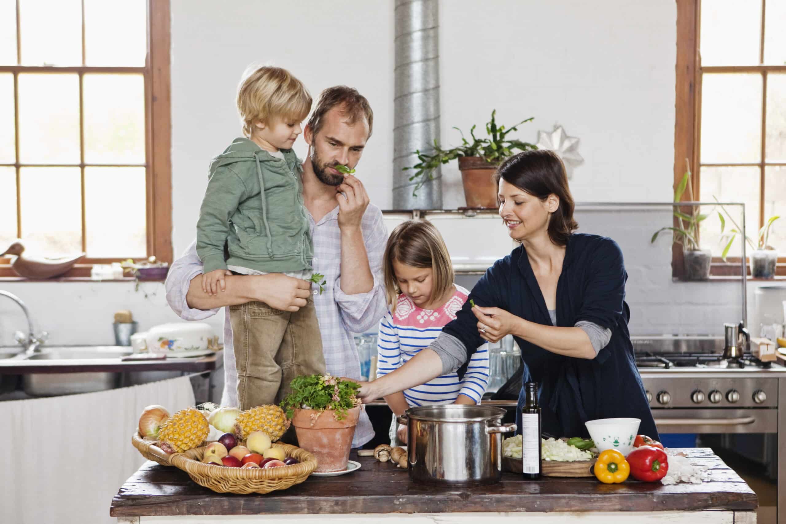 Eine vierköpfige Familie kocht gemeinsam in einer Küche. Ein Mann hält einen Jungen im Arm, und eine Frau und ein Mädchen bereiten auf einer Kücheninsel Essen mit Obst, Gemüse und Kräutern zu. Die Küche hat große Fenster, Topfpflanzen und Geräte aus Edelstahl. © Fotografie Tomas Rodriguez