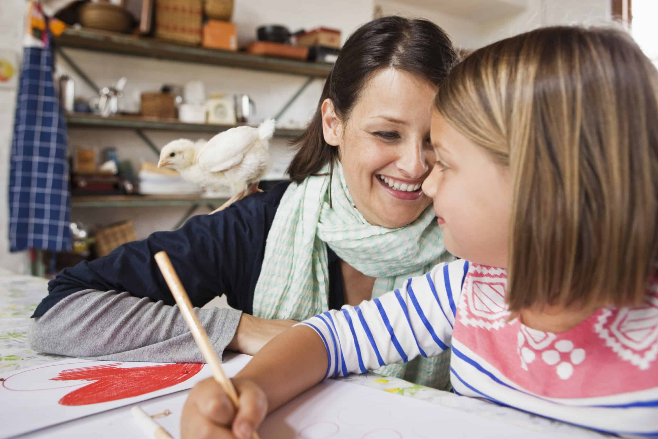 Eine Frau lächelt ein Kind herzlich an, das an einem Tisch malt. Die Frau trägt einen leichten Schal um den Hals und ein kleines Küken sitzt auf ihrer Schulter. Die Szene scheint eine gemütliche Küche mit Regalen im Hintergrund zu sein. © Fotografie Tomas Rodriguez
