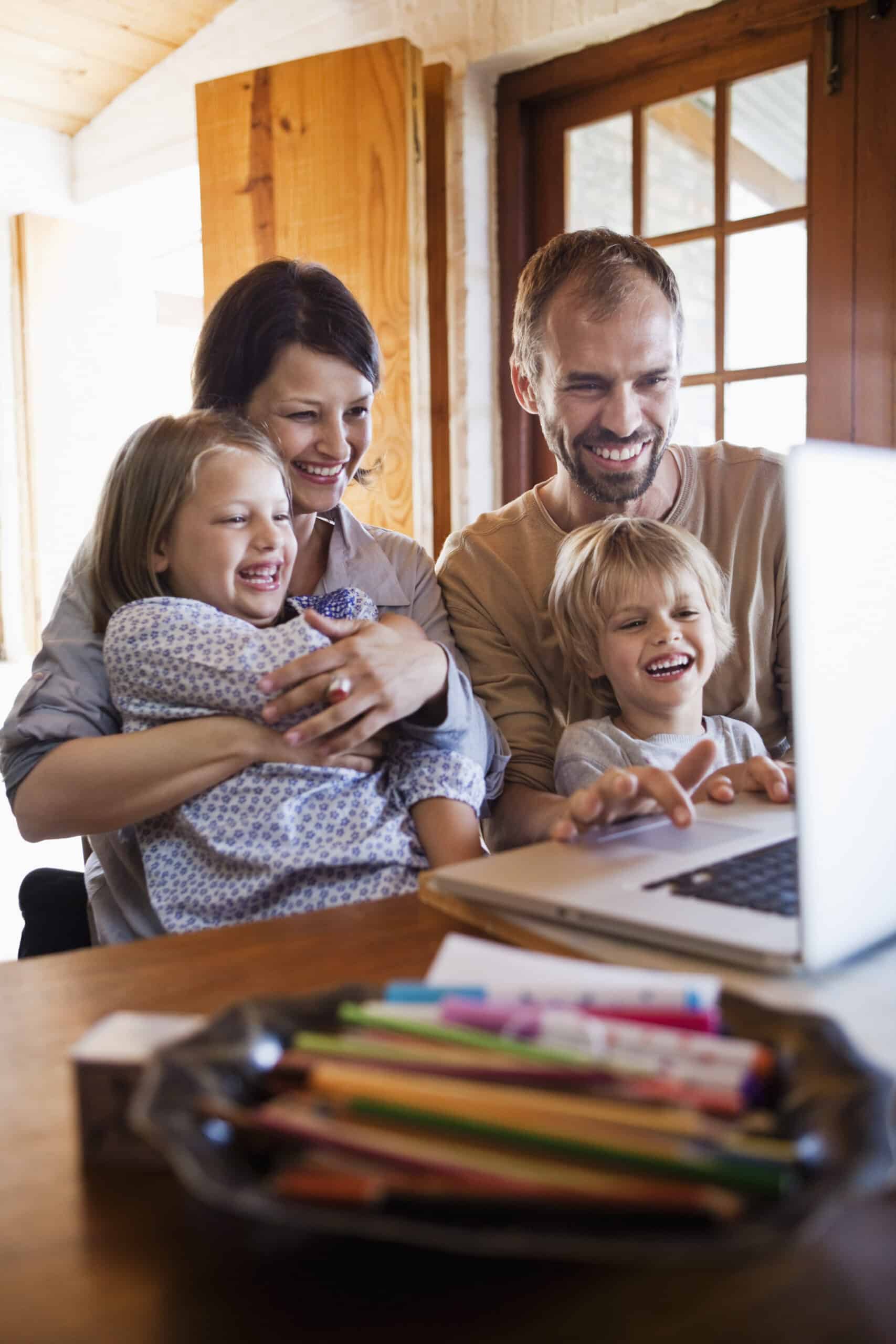 Eine glückliche vierköpfige Familie, darunter zwei Kinder, sitzt an einem Holztisch um einen Laptop herum. Sie lächeln und lachen, während sie auf den Bildschirm schauen. Auf dem Tisch steht eine Schüssel voller Buntstifte und anderer Malutensilien. Der Raum ist hell und hat natürliches Licht. © Fotografie Tomas Rodriguez