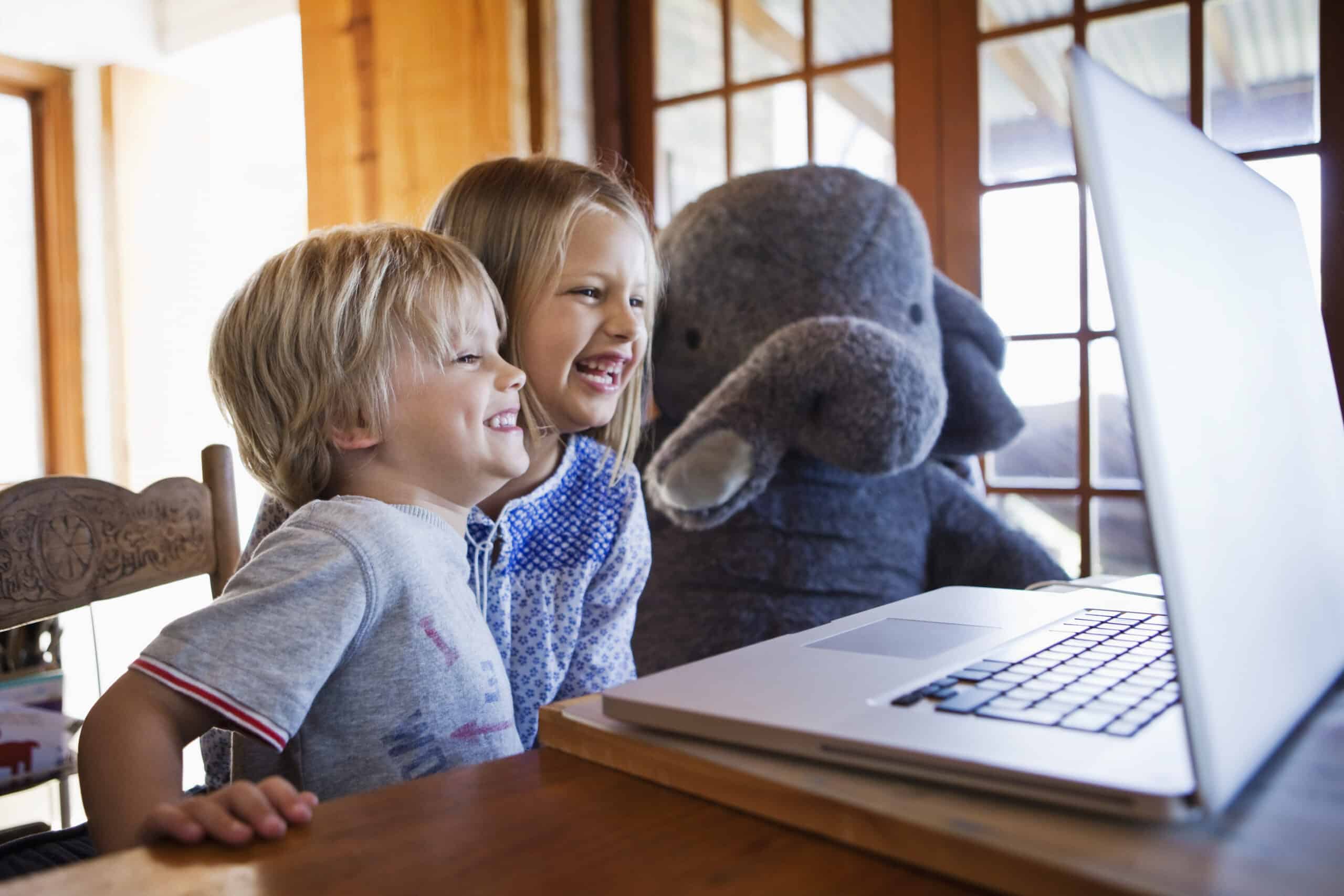Zwei lächelnde Kinder sitzen an einem Tisch mit einem großen Stoffelefanten und schauen auf einen Laptop-Bildschirm. Der Raum ist mit Holzakzenten warm beleuchtet und große Fenster im Hintergrund lassen natürliches Licht herein. © Fotografie Tomas Rodriguez