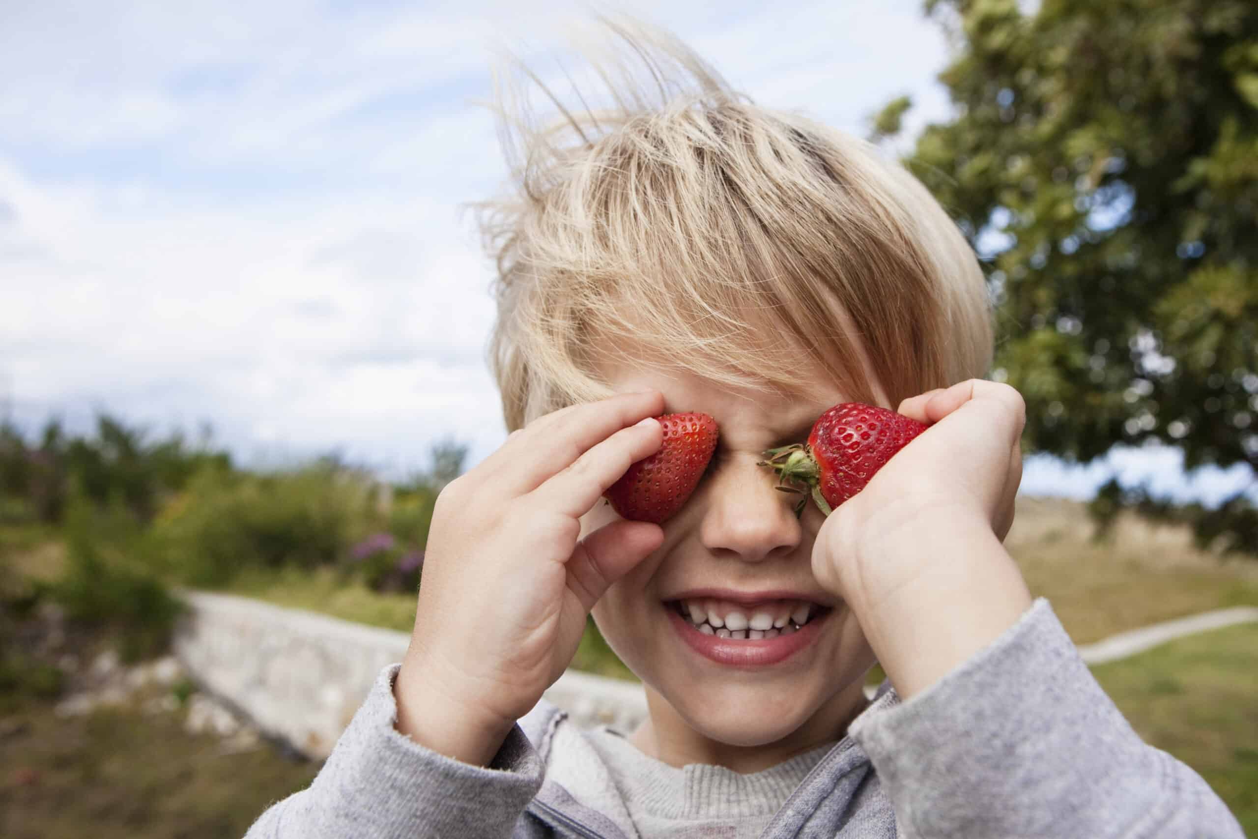 Ein kleines Kind mit blondem Haar hält sich spielerisch zwei Erdbeeren vor die Augen und lächelt breit. Das Kind ist an einem windigen Tag im Freien, im Hintergrund sind Grünflächen und ein Weg zu sehen. © Fotografie Tomas Rodriguez