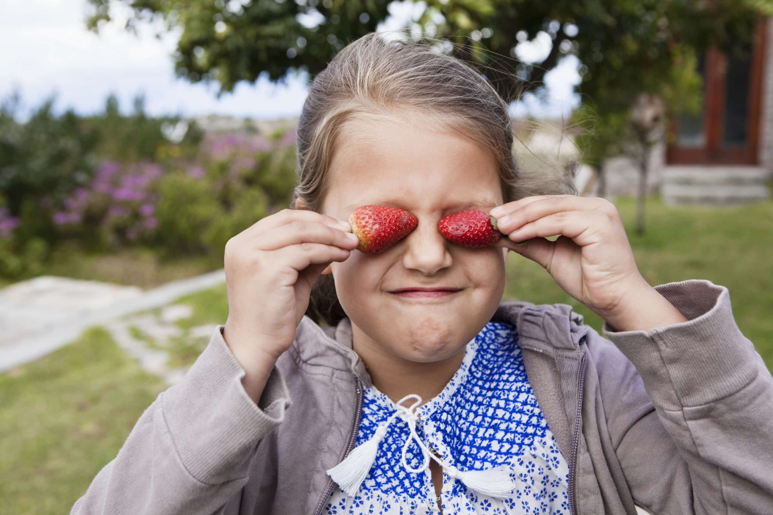 Ein junges Mädchen steht mit verspieltem Gesichtsausdruck im Freien und hält zwei Erdbeeren vor die Augen. Sie trägt ein blau-weiß gemustertes Oberteil unter einer leichten Jacke, im Hintergrund sind verschwommenes Grün und Blumen zu sehen. © Fotografie Tomas Rodriguez
