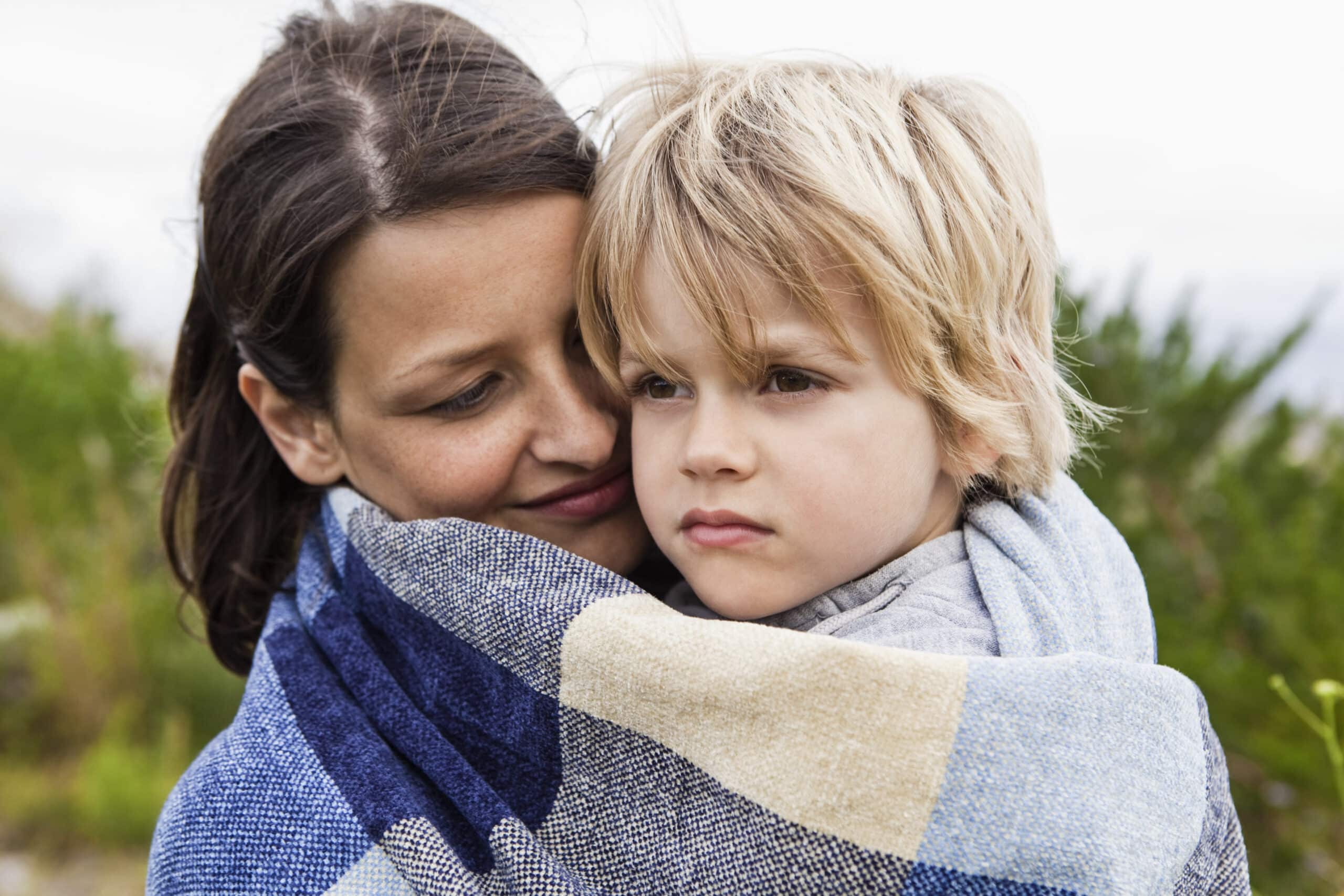 Eine Frau und ein kleines Kind sind in eine große blau-beige karierte Decke gehüllt. Die Frau hat dunkles Haar und schmiegt sich an den Kopf des Kindes, während das Kind mit blondem Haar in die Ferne blickt. Sie sind draußen mit viel Grün im Hintergrund. © Fotografie Tomas Rodriguez