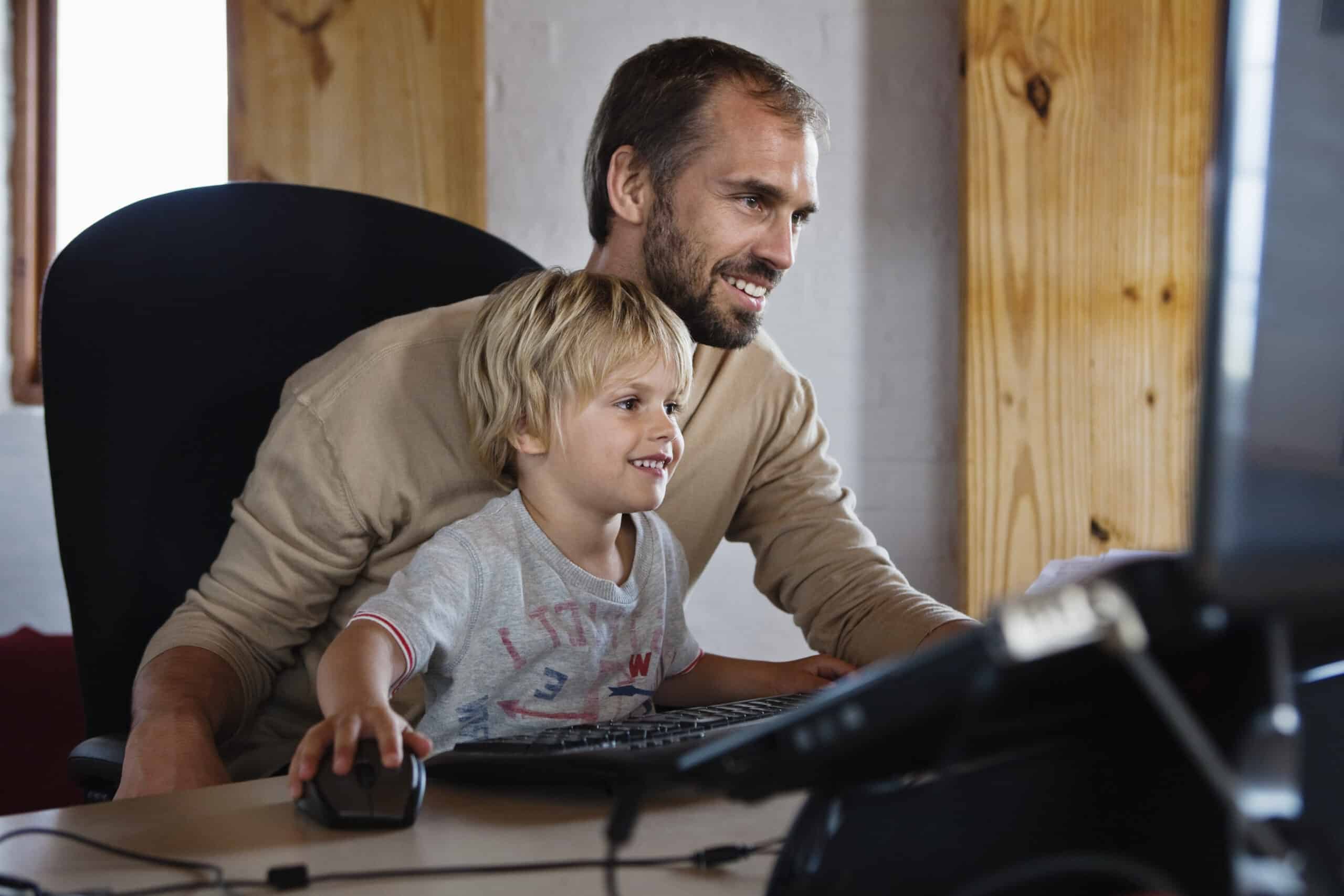 Ein Mann und ein kleines Kind sitzen an einem Schreibtisch. Beide lächeln und schauen auf einen Computerbildschirm. Das Kind benutzt die Maus, während der Mann hinter ihm sitzt und es mit einer Hand an der Maus und der anderen Hand an der Tastatur führt. Der Raum ist mit Holzakzenten versehen. © Fotografie Tomas Rodriguez