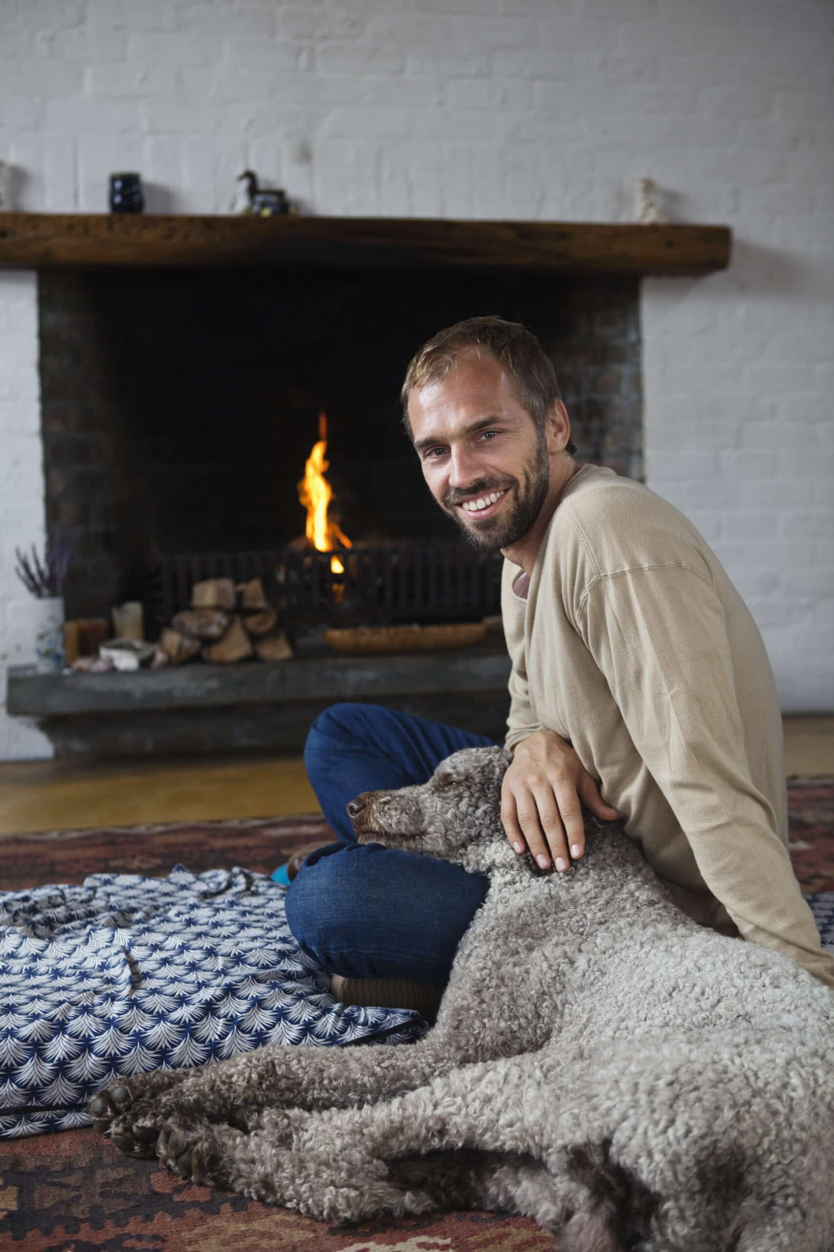 Eine Person mit Bart und beigem Pullover sitzt auf einem gemusterten Teppich vor einem brennenden Kamin und lächelt, während sie einen braunen, lockigen Hund streichelt, der neben ihr liegt. Im Kamin ist ein Stapel Brennholz zu sehen. Der Raum hat eine gemütliche Atmosphäre. © Fotografie Tomas Rodriguez