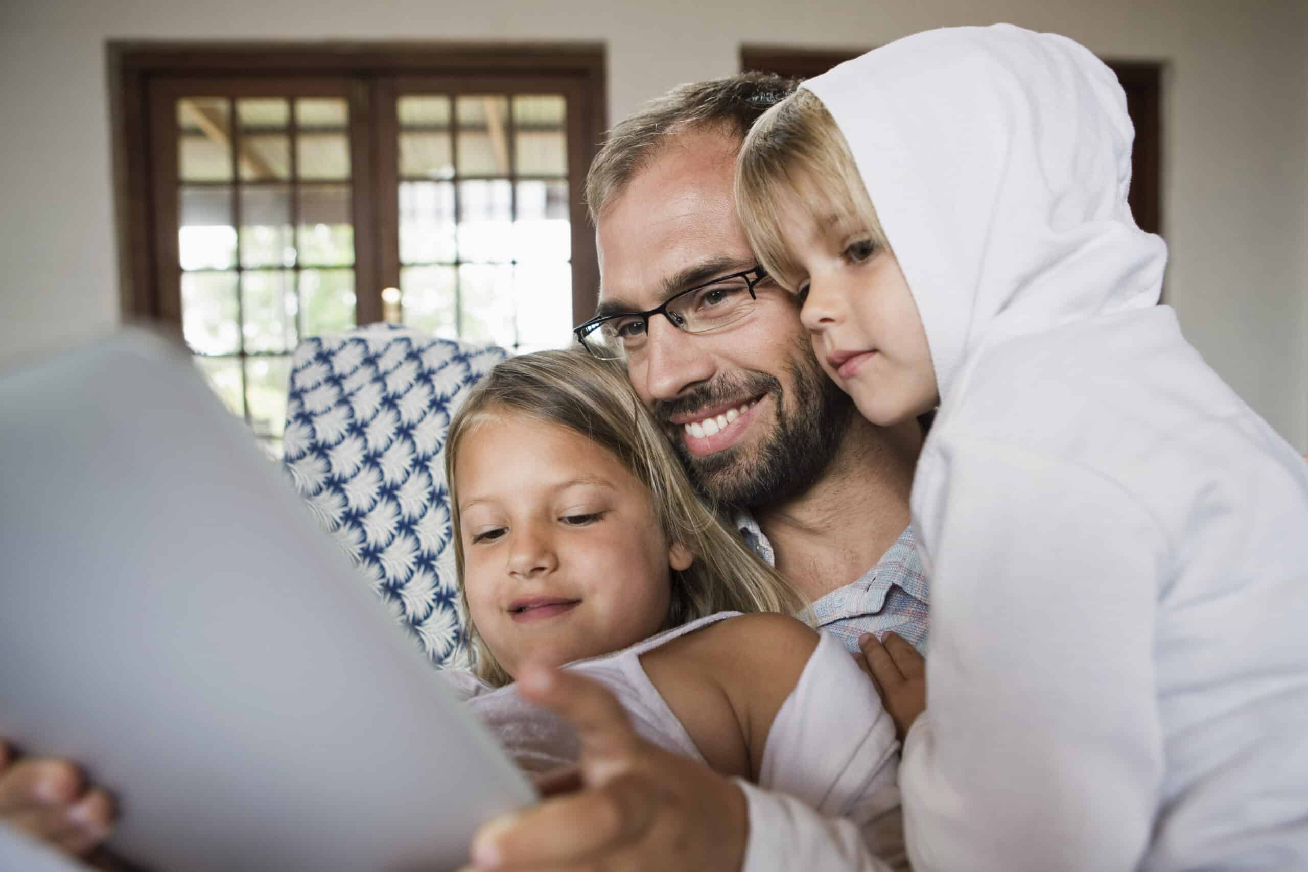 Ein Mann mit Brille und Bart, der ein hellblaues Hemd trägt, sitzt mit zwei Kindern auf einem Sofa. Ein Kind trägt einen weißen Kapuzenpullover und das andere ein weißes Tanktop. Sie lächeln alle und schauen gemeinsam auf einen Tablet-Bildschirm. © Fotografie Tomas Rodriguez