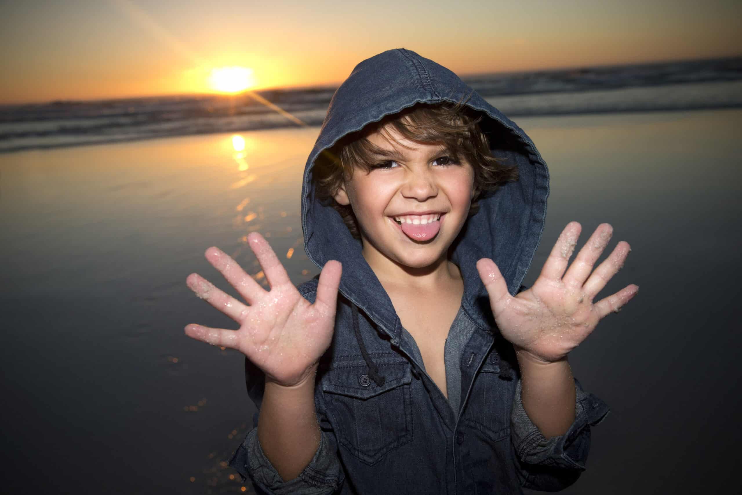 Ein verspieltes Kind in einer Jeansjacke mit Kapuze steht bei Sonnenuntergang an einem Strand. Das Kind lächelt breit, streckt die Zunge heraus und hält sandige Hände hoch, während im Hintergrund die Sonne über dem Meer untergeht. © Fotografie Tomas Rodriguez