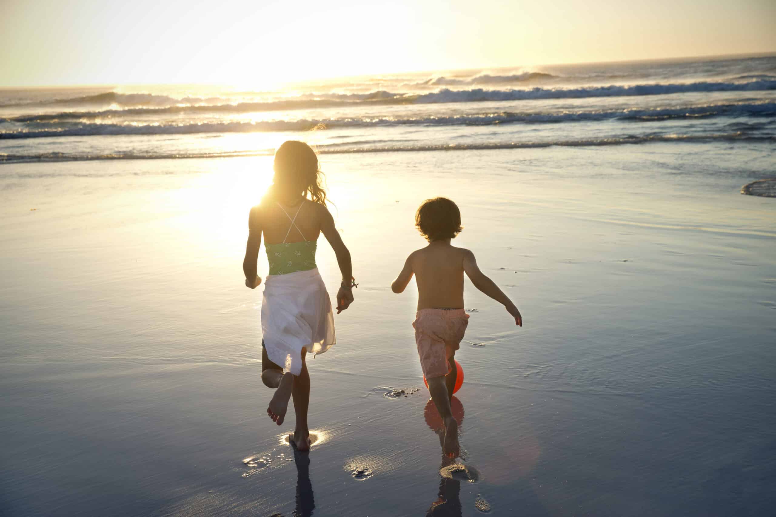 Zwei Kinder, ein Mädchen und ein Junge, laufen bei Sonnenuntergang barfuß am Ufer entlang. Die Sonne geht hinter ihnen unter und wirft lange Schatten auf den Sand. Das Mädchen trägt ein Top und einen weißen Rock, während der Junge nur Shorts trägt. Im Hintergrund brechen sanft die Wellen. © Fotografie Tomas Rodriguez