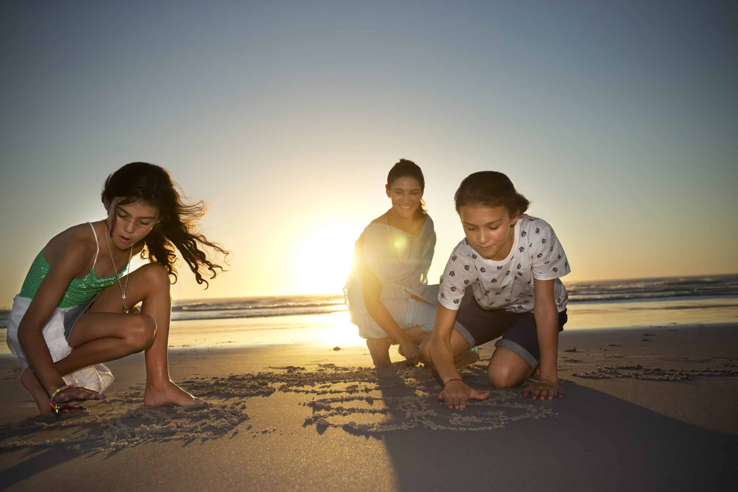 Drei Kinder spielen bei Sonnenuntergang im Sand am Strand. Ein Kind links malt mit einem Stock, das mittlere Kind lächelt und das Kind rechts malt ebenfalls in den Sand. Hinter ihnen geht die Sonne unter und wirft lange Schatten auf den Sand. © Fotografie Tomas Rodriguez