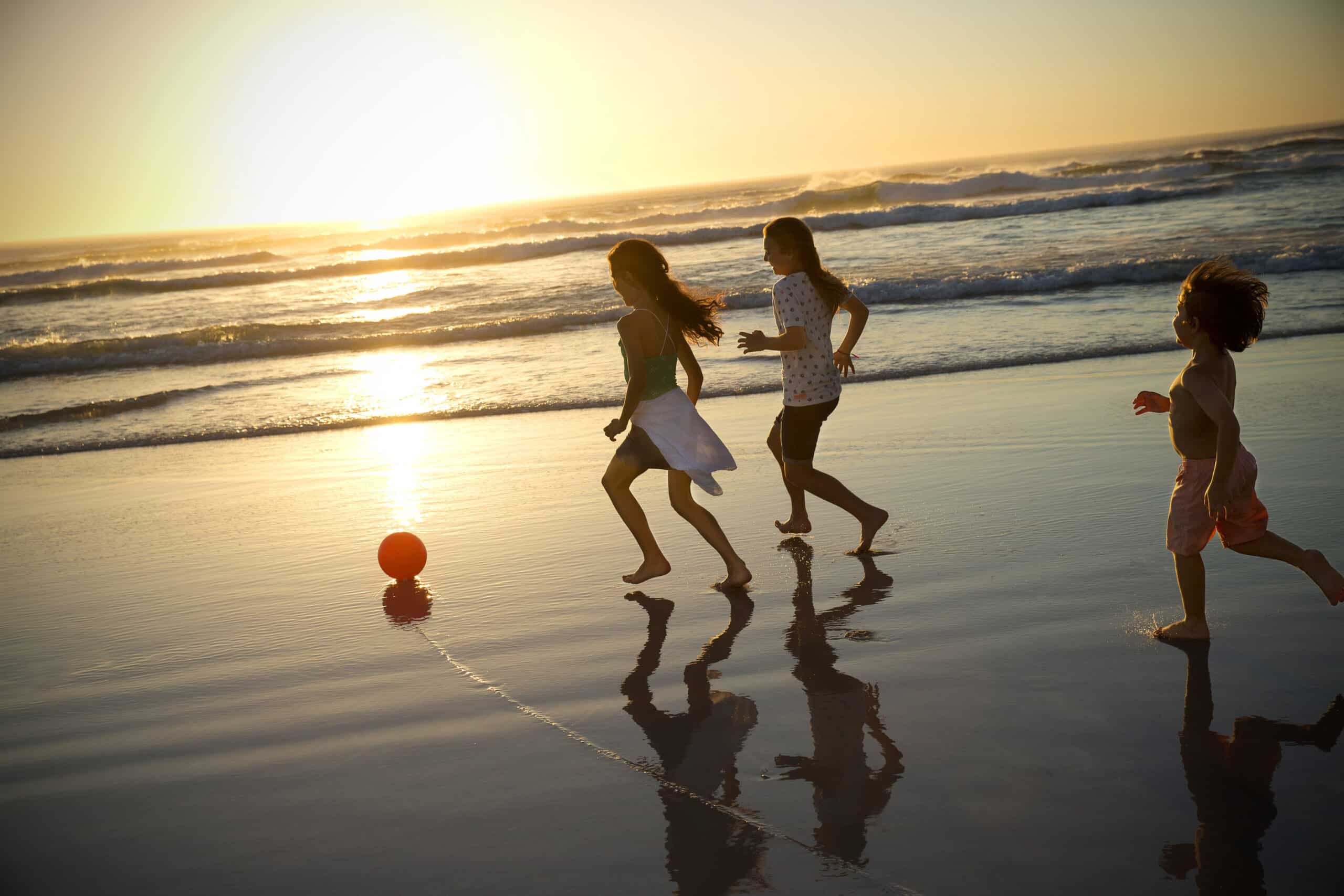 Drei Kinder spielen bei Sonnenuntergang mit einem roten Ball an einem Strand. Sie laufen auf dem nassen Sand in der Nähe des Wasserrands. Die Sonne steht tief am Horizont, wirft ein warmes goldenes Licht und erzeugt lange Schatten. Im Hintergrund sind Wellen zu sehen. © Fotografie Tomas Rodriguez
