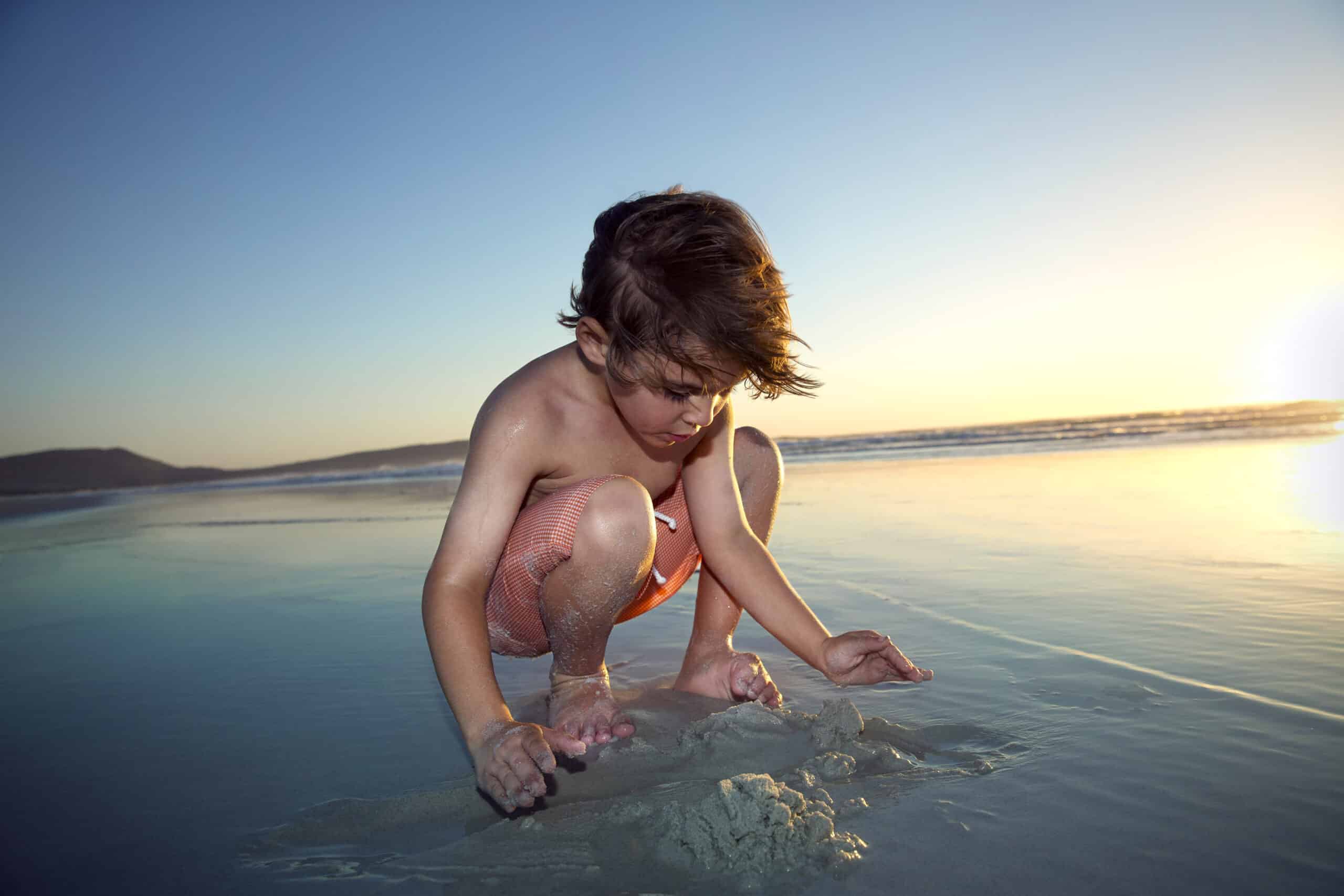 Ein kleines Kind mit nassen Haaren und orangefarbener Badehose hockt bei Sonnenuntergang im seichten Wasser am Strand und schaut aufmerksam auf den Sand, mit dem es spielt. Im Hintergrund sind der Horizont und sanfte Wellen unter einem klaren Himmel sichtbar. © Fotografie Tomas Rodriguez