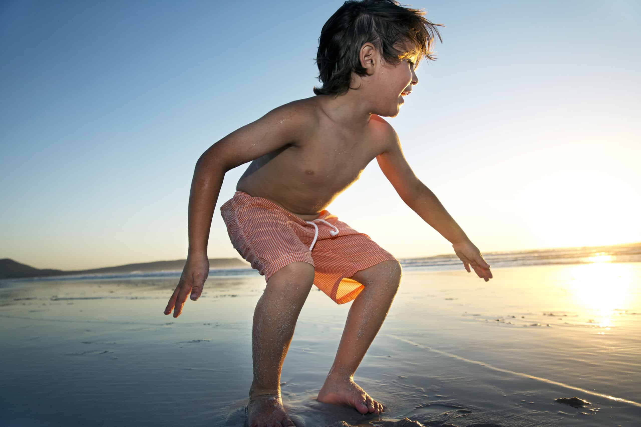Ein kleiner Junge in orangefarbener Badehose steht mit gebeugten Knien und ausgestreckten Armen an einem nassen Strand und blickt zum Horizont. Die Sonne steht tief am Himmel und wirft einen warmen Schein und lange Schatten auf den Sand. Im Hintergrund plätschern sanft Wellen ans Ufer. © Fotografie Tomas Rodriguez