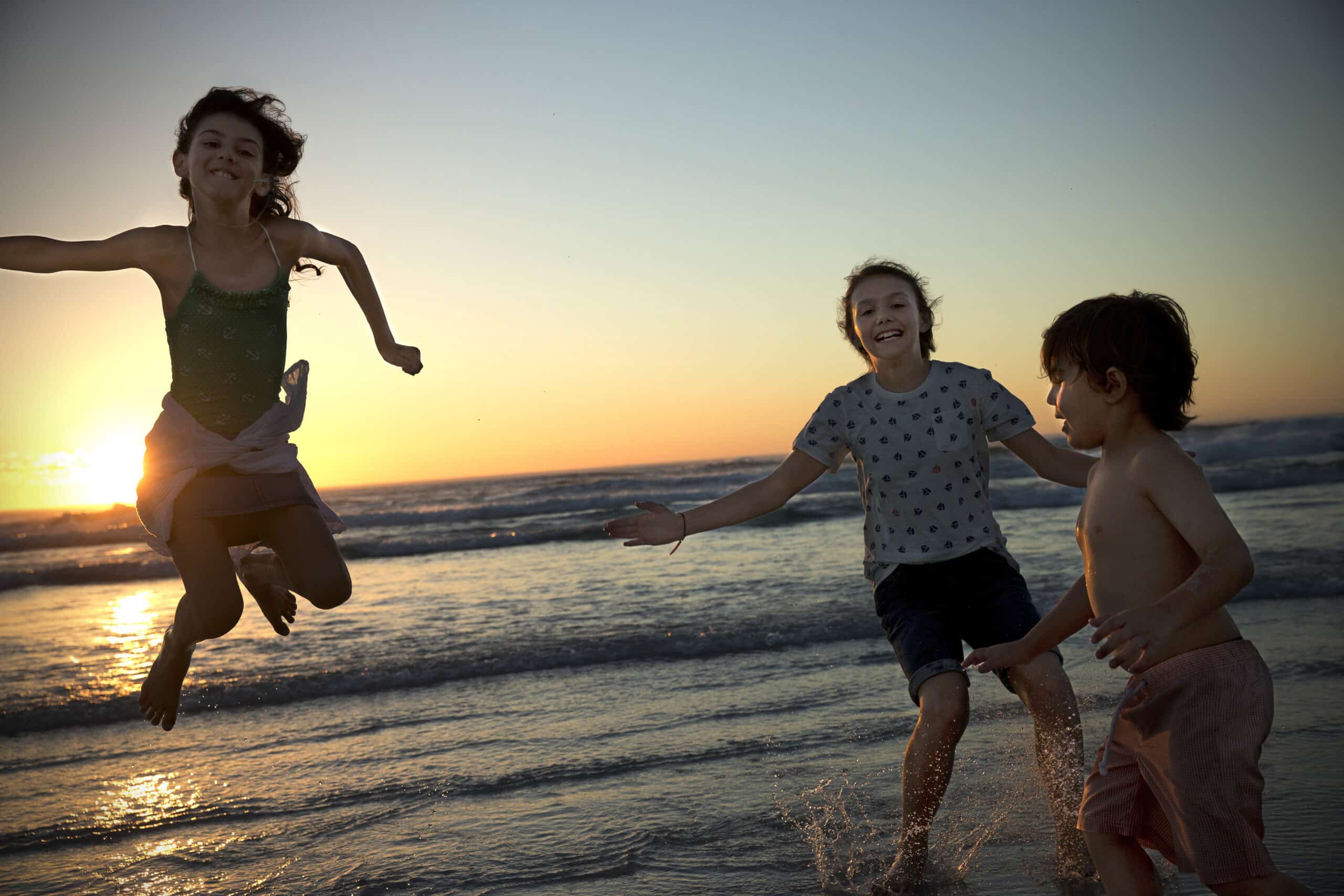 Drei Kinder spielen fröhlich in den seichten Wellen am Strand bei Sonnenuntergang. Ein Mädchen springt in die Luft, ein anderes lächelt und ein Junge ohne Hemd planscht im Wasser. Der Himmel ist in einem sanften Farbverlauf aus warmen Farben gehalten, während die Sonne am Horizont untergeht. © Fotografie Tomas Rodriguez