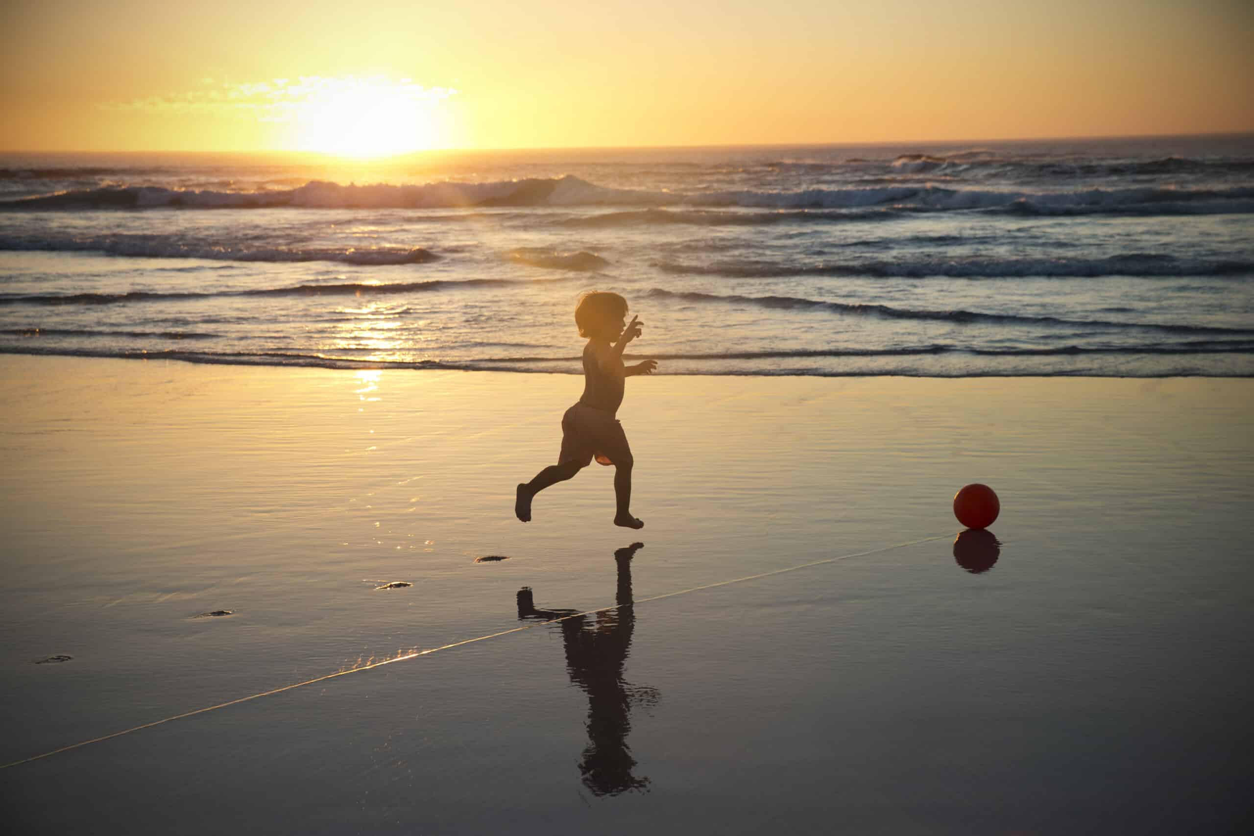 Ein kleines Kind rennt während eines leuchtenden Sonnenuntergangs auf einen roten Ball zu. Im Hintergrund rollen sanft die Meereswellen und der nasse Sand spiegelt das Kind, den Ball und die goldenen Farbtöne der Sonne wider. © Fotografie Tomas Rodriguez