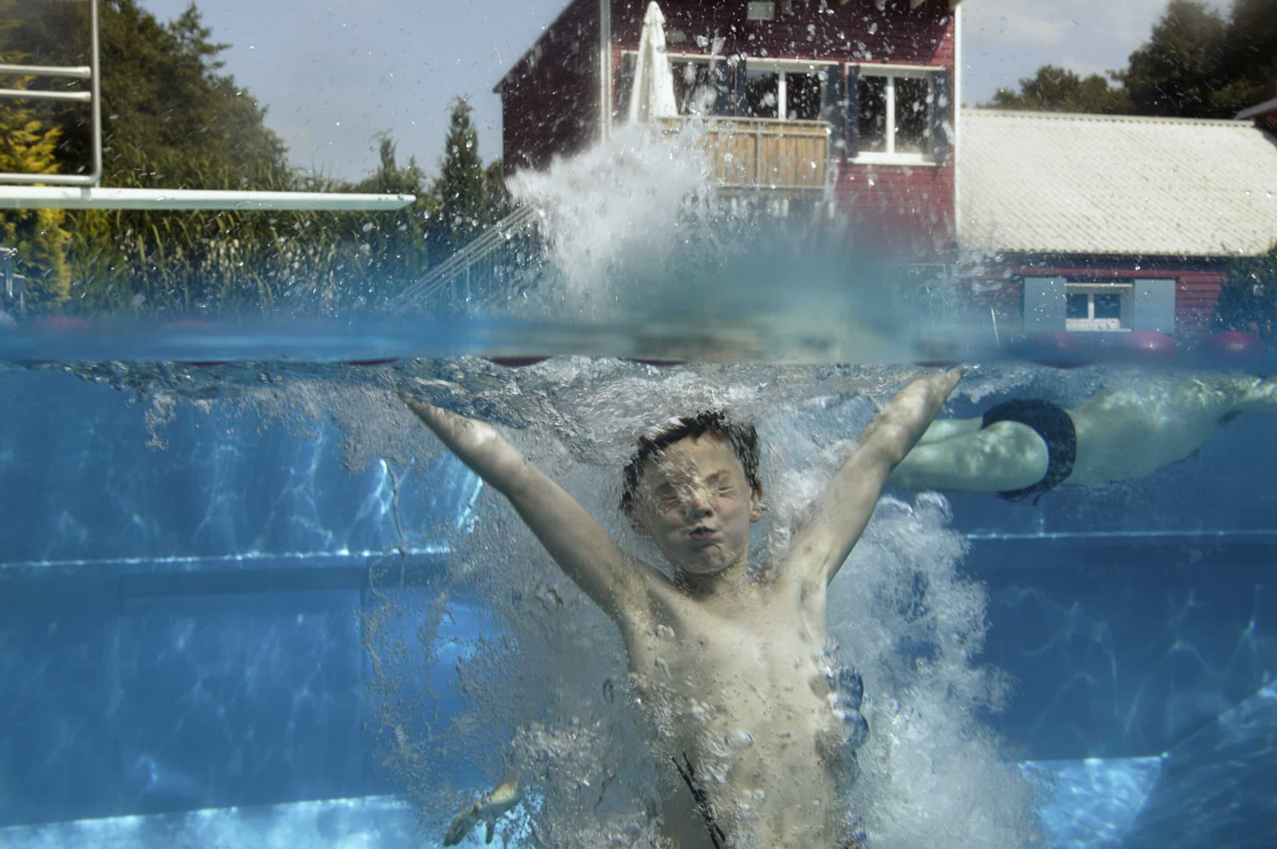 Eine Person ist in einem Schwimmbecken untergetaucht und mitten im Planschen mit ausgestreckten Armen und geschlossenen Augen aufgenommen. Im Hintergrund sind verschwommene rot-weiße Gebäude über dem Wasser zu sehen. Die Sonne scheint und schafft eine helle und lebendige Atmosphäre. © Fotografie Tomas Rodriguez