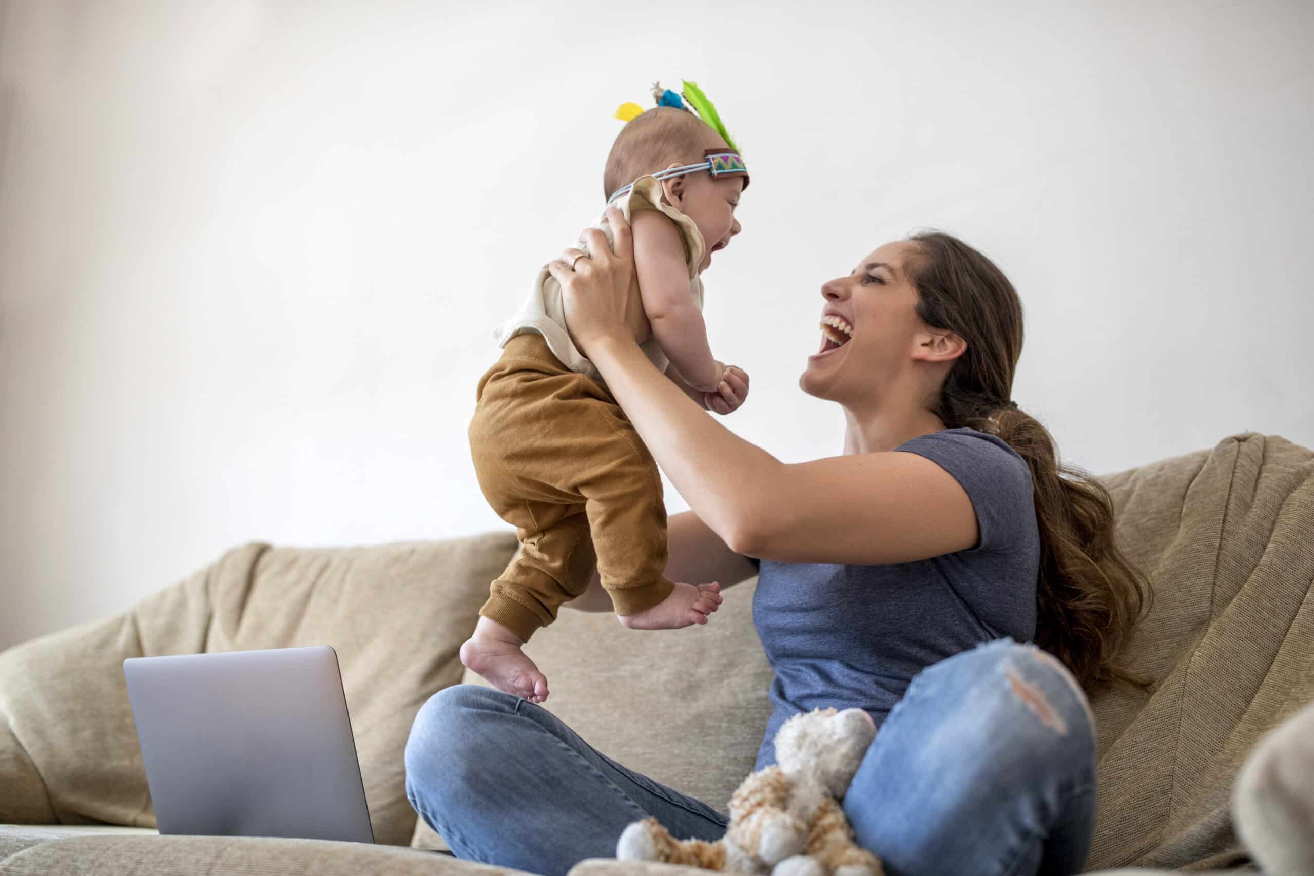 Eine Frau sitzt auf einem Sofa und hebt mit freudigem Gesichtsausdruck ein Baby in die Luft. Das Baby trägt eine braune Hose und ein buntes Stirnband. Ein Laptop und ein Stofftier liegen neben ihnen auf dem Sofa. © Fotografie Tomas Rodriguez