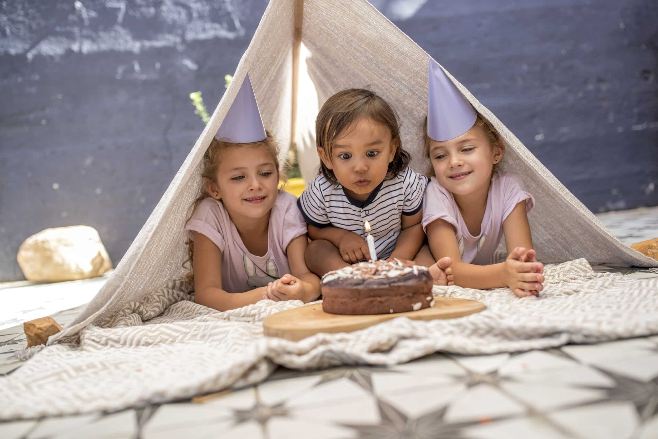 Drei Kinder mit Partyhüten liegen auf dem Bauch unter einem Stoffzelt im Freien und blicken gespannt auf einen Schokoladenkuchen auf einem Holzbrett vor ihnen. Der Boden ist mit einem geometrischen Muster versehen, im Hintergrund ist ein Stein zu sehen. © Fotografie Tomas Rodriguez