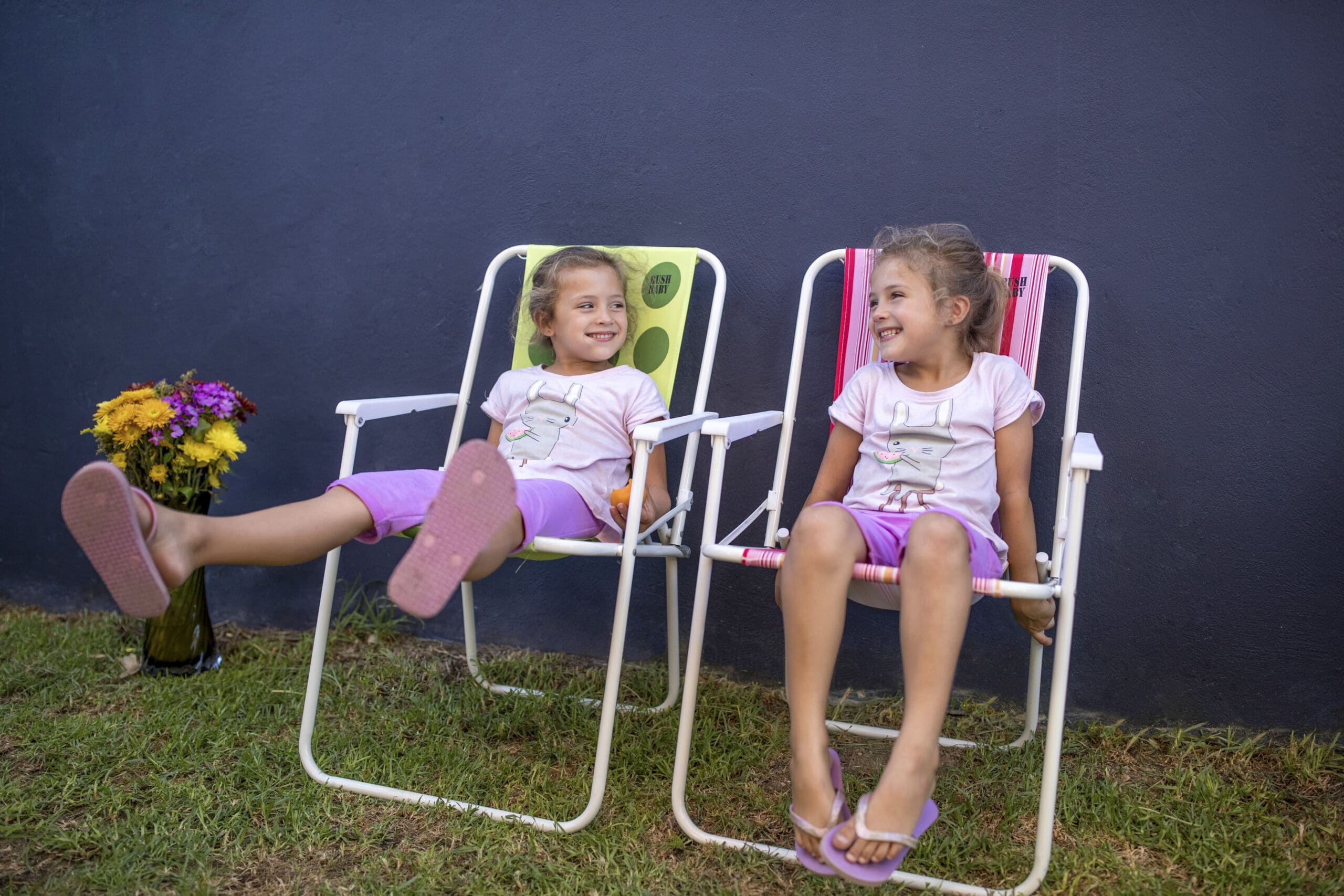 Zwei junge Mädchen, in passenden Outfits aus rosa Oberteilen und lila Shorts, sitzen auf Gartenstühlen vor einer dunkelgrauen Wand. Beide lächeln und lachen. Ein Mädchen streckt die Beine aus, während das andere im Schneidersitz sitzt. Neben ihnen steht eine Vase mit bunten Blumen. © Fotografie Tomas Rodriguez