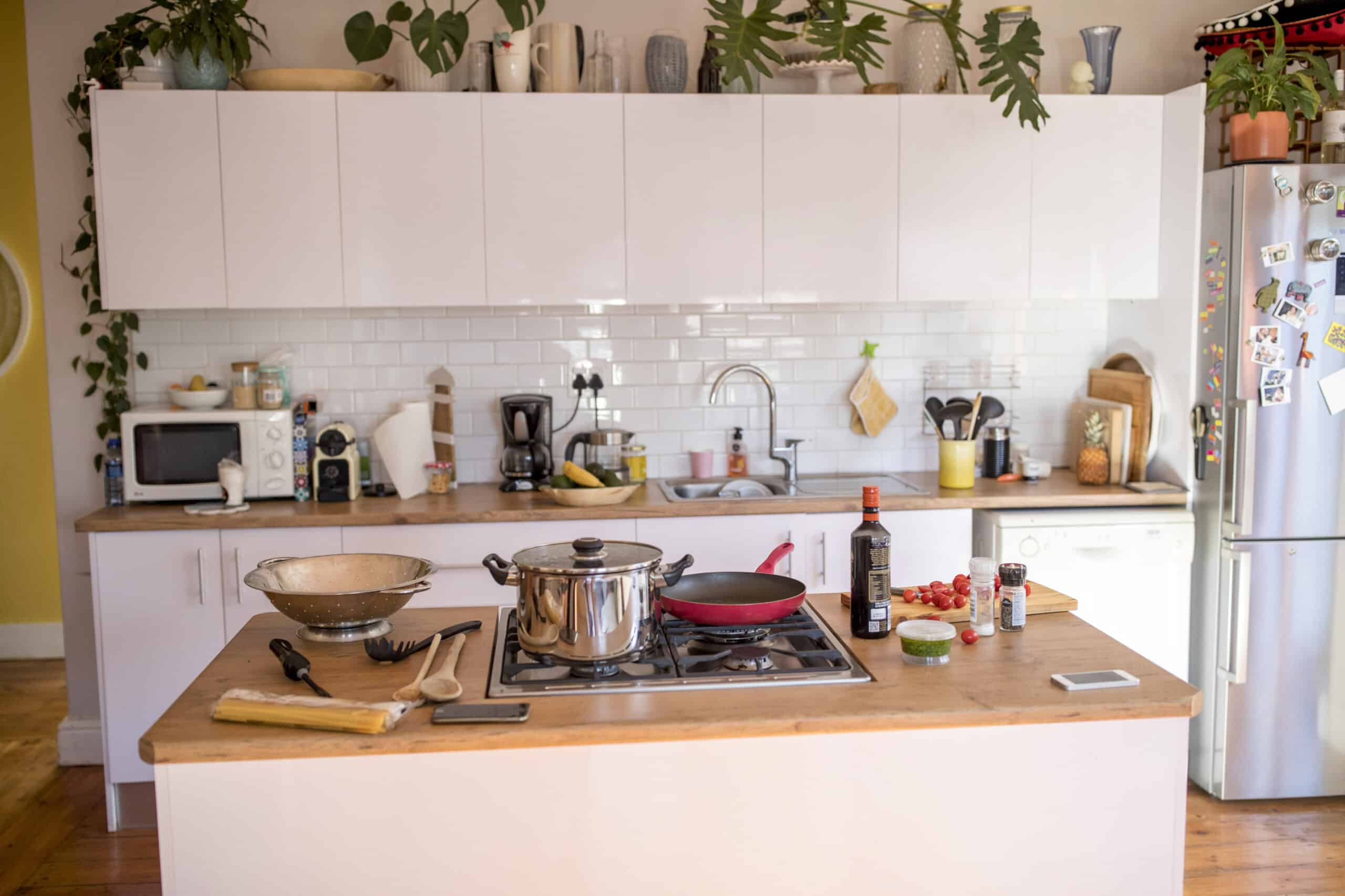 Eine Küche mit einer Holzarbeitsplatte, auf der ein Topf, eine Bratpfanne, Gemüse und eine Flasche Kochwein stehen. Im Hintergrund sind weiße Schränke, eine Mikrowelle, eine Kaffeemaschine, Pflanzen und ein mit Magneten dekorierter Kühlschrank zu sehen. © Fotografie Tomas Rodriguez