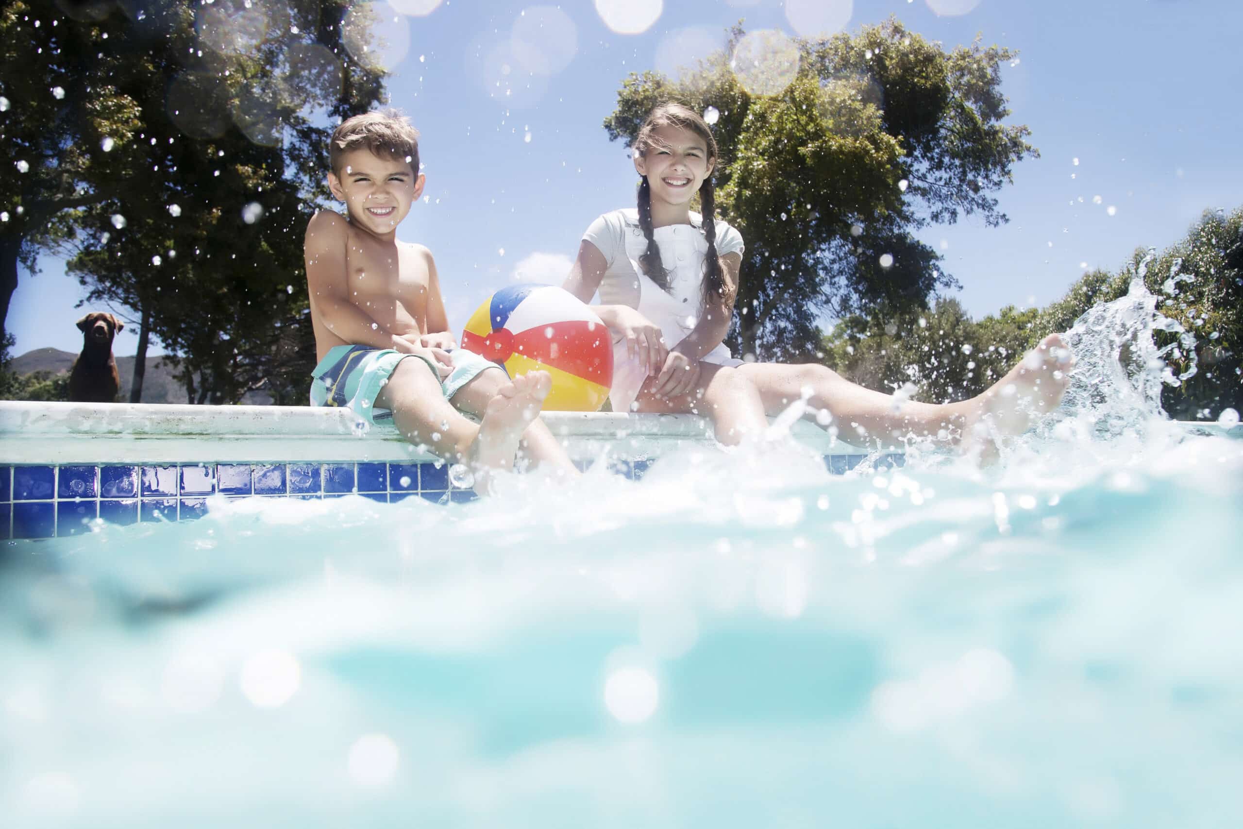 Zwei Kinder sitzen am Rand eines Schwimmbeckens, lächeln und plantschen mit ihren Füßen im Wasser. Ein Kind hält einen bunten Wasserball. Im Hintergrund sind Bäume zu sehen, und es ist ein sonniger Tag mit klarem, blauem Himmel. © Fotografie Tomas Rodriguez
