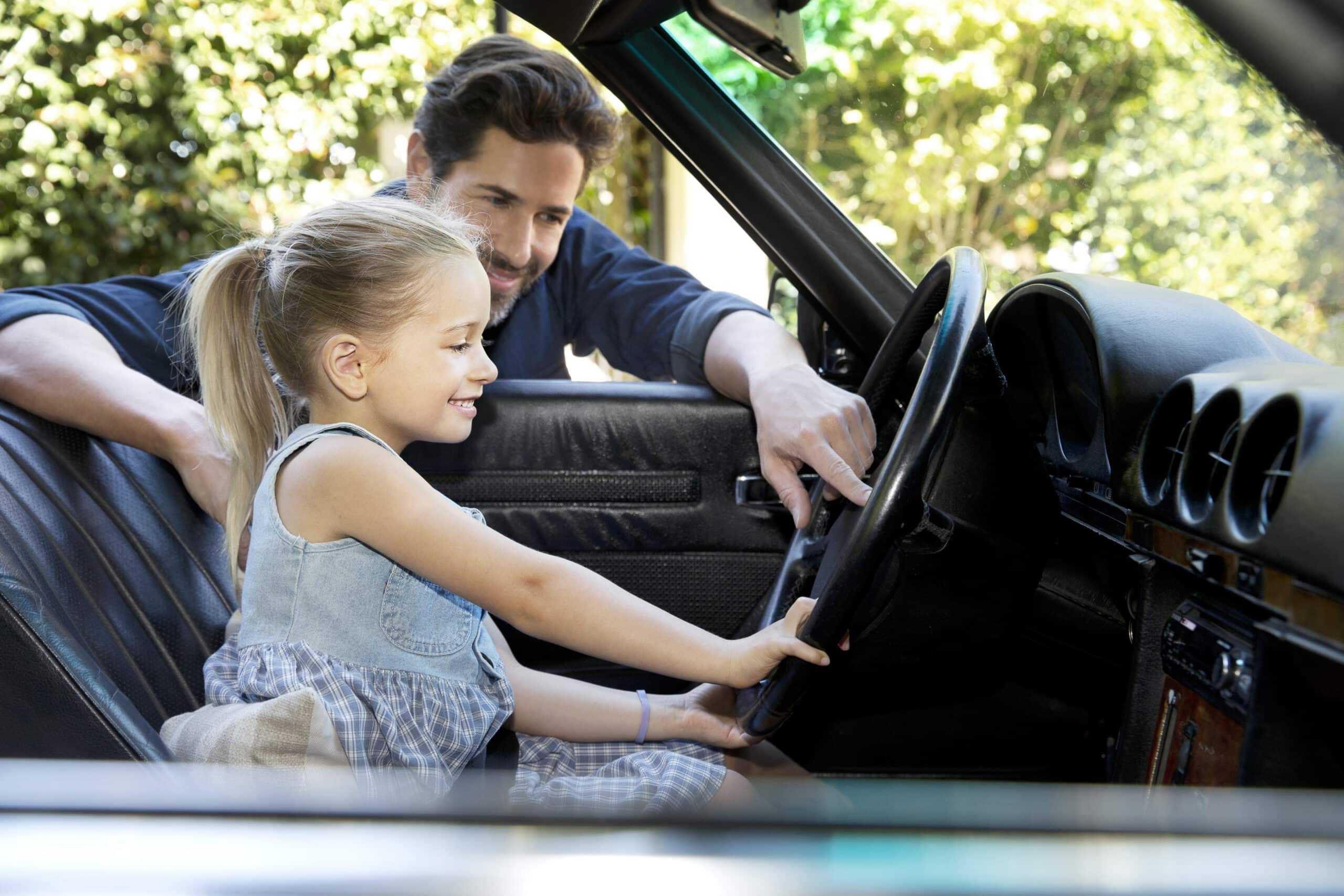 Ein lächelndes junges Mädchen mit blondem Haar und einem blauen Kleid sitzt auf dem Fahrersitz eines Autos und hält das Lenkrad in der Hand. Ein Mann mit dunklem Haar steht außerhalb des Autos, lehnt sich durch die offene Tür hinein, lächelt und beobachtet sie. Im Hintergrund sind Bäume zu sehen. © Fotografie Tomas Rodriguez