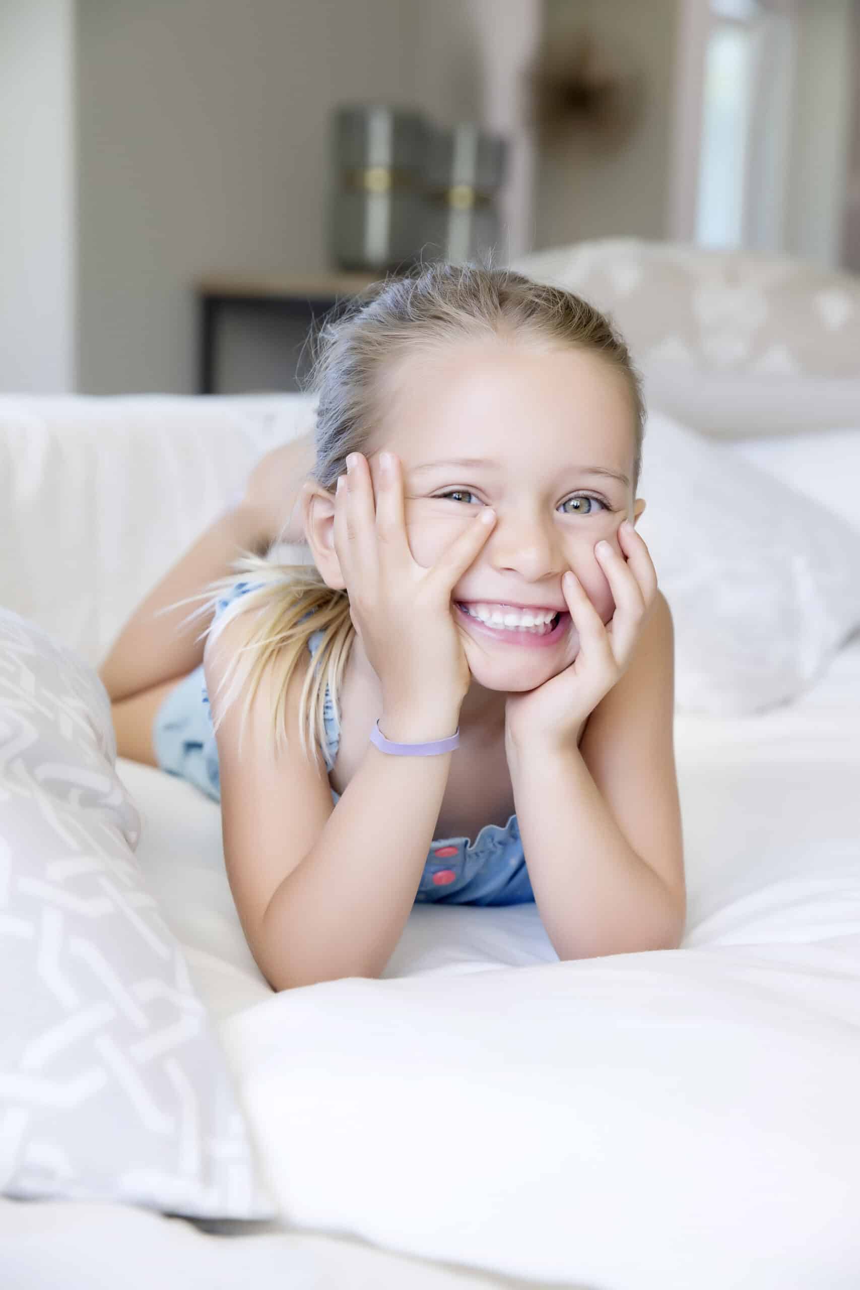 Ein kleines Kind mit blonden Haaren und einem blauen ärmellosen Kleid liegt auf einem Bett, auf die Ellbogen gestützt, lächelt breit und hält sein Gesicht mit beiden Händen. Die Bettwäsche ist weiß und gemustert. Der Hintergrund ist leicht verschwommen. © Fotografie Tomas Rodriguez