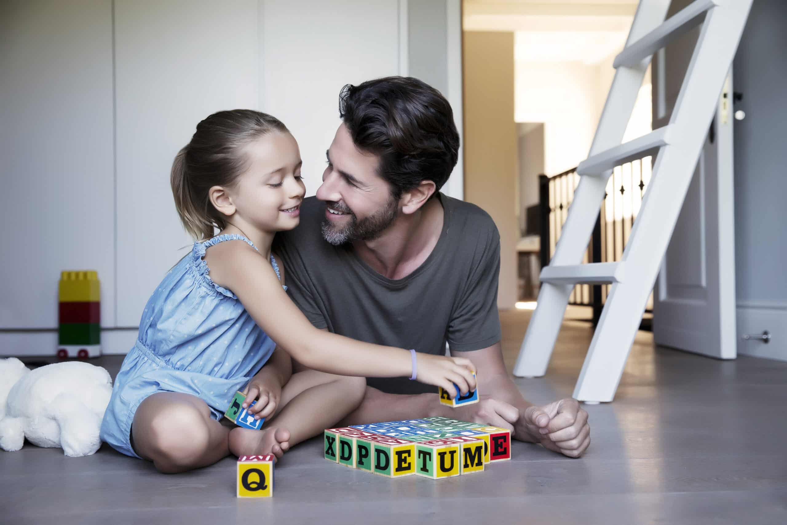 Ein junges Mädchen und ein Mann sitzen in einem hell erleuchteten Raum auf dem Boden, lächeln einander an und spielen mit bunten Alphabetblöcken. Das Mädchen trägt ein blaues Kleid und der Mann ein graues T-Shirt. Neben ihnen liegt auf dem Boden ein weißes Stofftier. © Fotografie Tomas Rodriguez