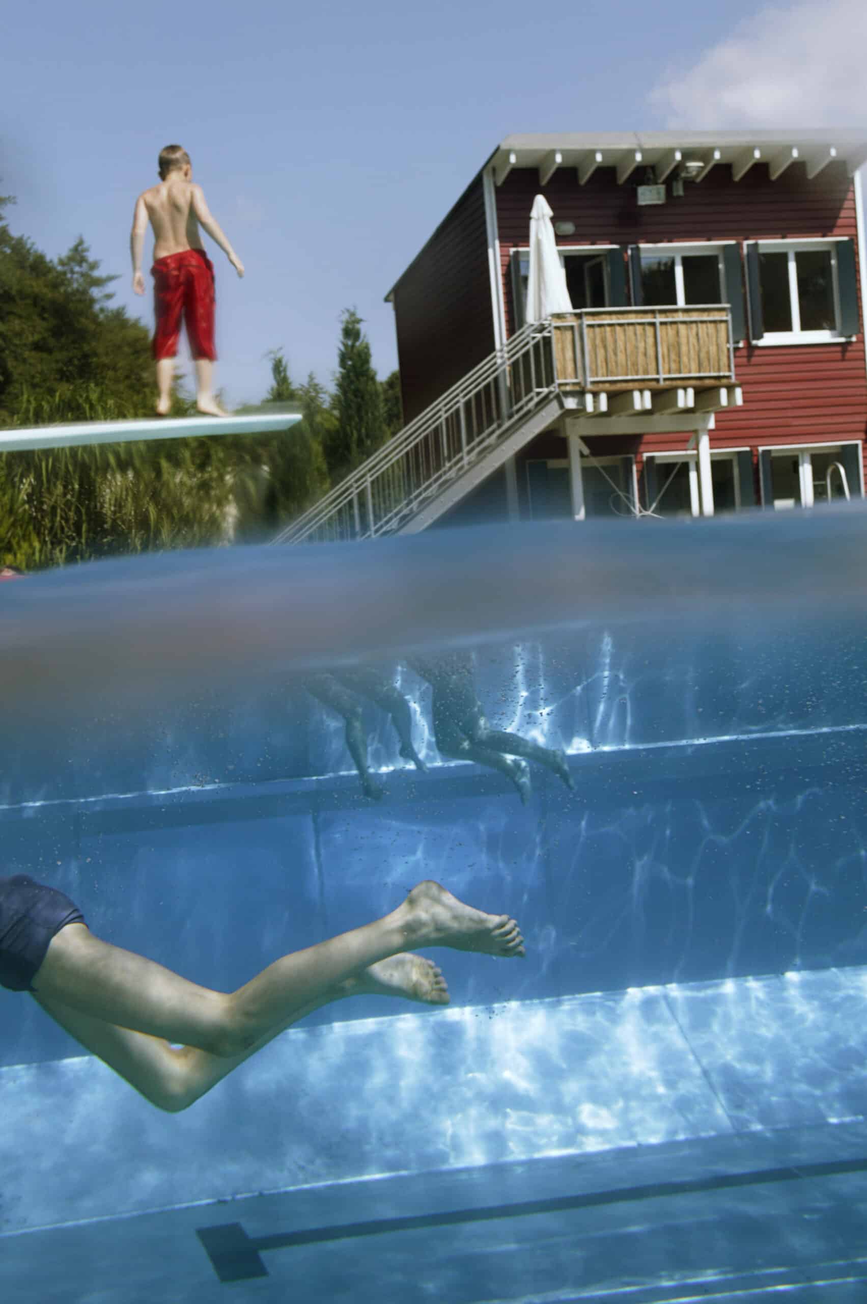 Ein teilweise untergetauchtes Bild zeigt einen Jungen in roter Badehose, der auf einem Sprungbrett über dem Wasser steht, während unter Wasser eine andere Person in einem Pool schwimmt. Im Hintergrund ist ein zweistöckiges Haus mit Balkon und Treppe zu sehen. © Fotografie Tomas Rodriguez