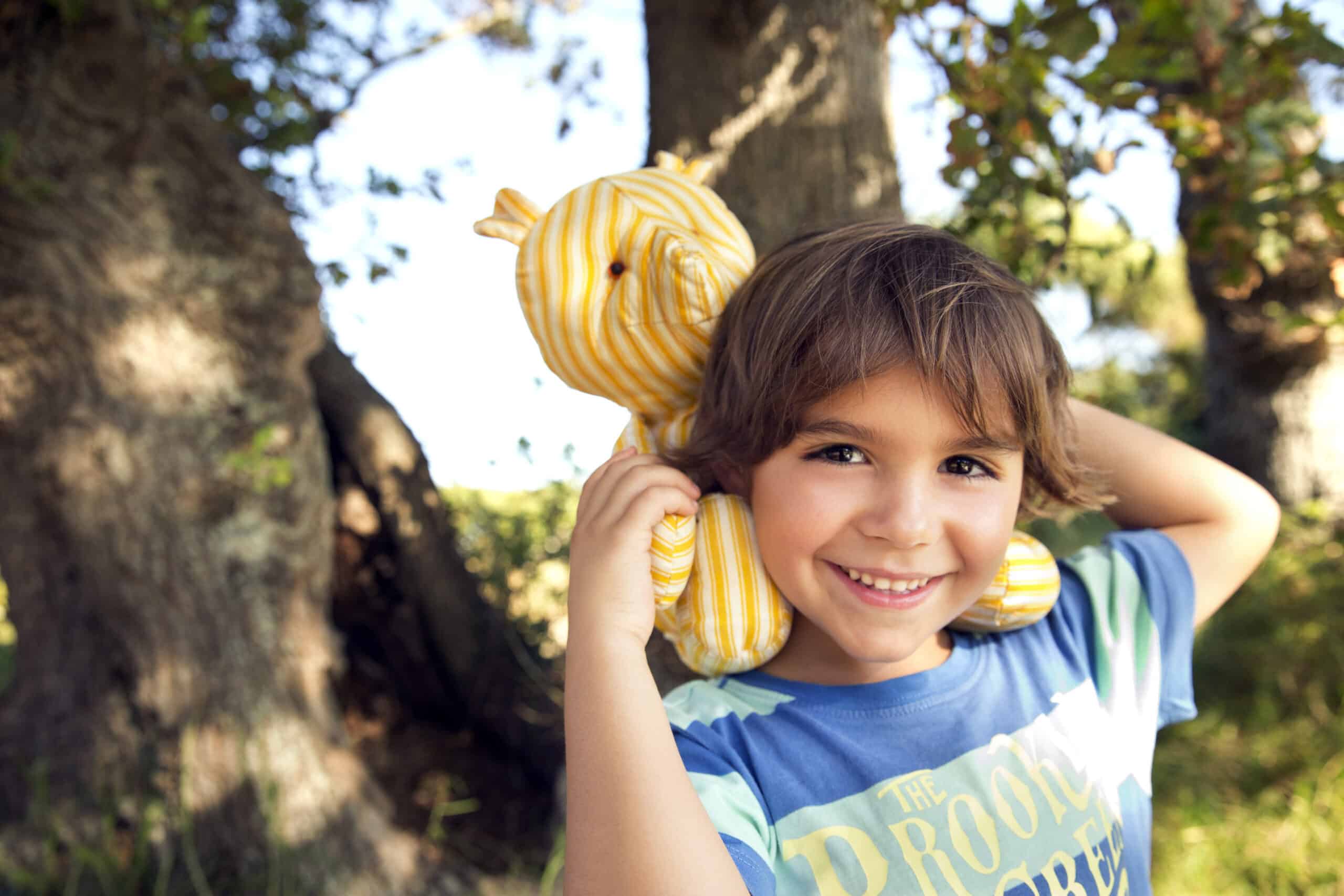 Ein lächelndes kleines Kind mit braunem Haar steht draußen vor Bäumen und hält ein gelb-weiß gestreiftes Stofftier über den Schultern. Das Kind trägt ein blau-grünes T-Shirt und der Hintergrund ist sonnig und grün. © Fotografie Tomas Rodriguez