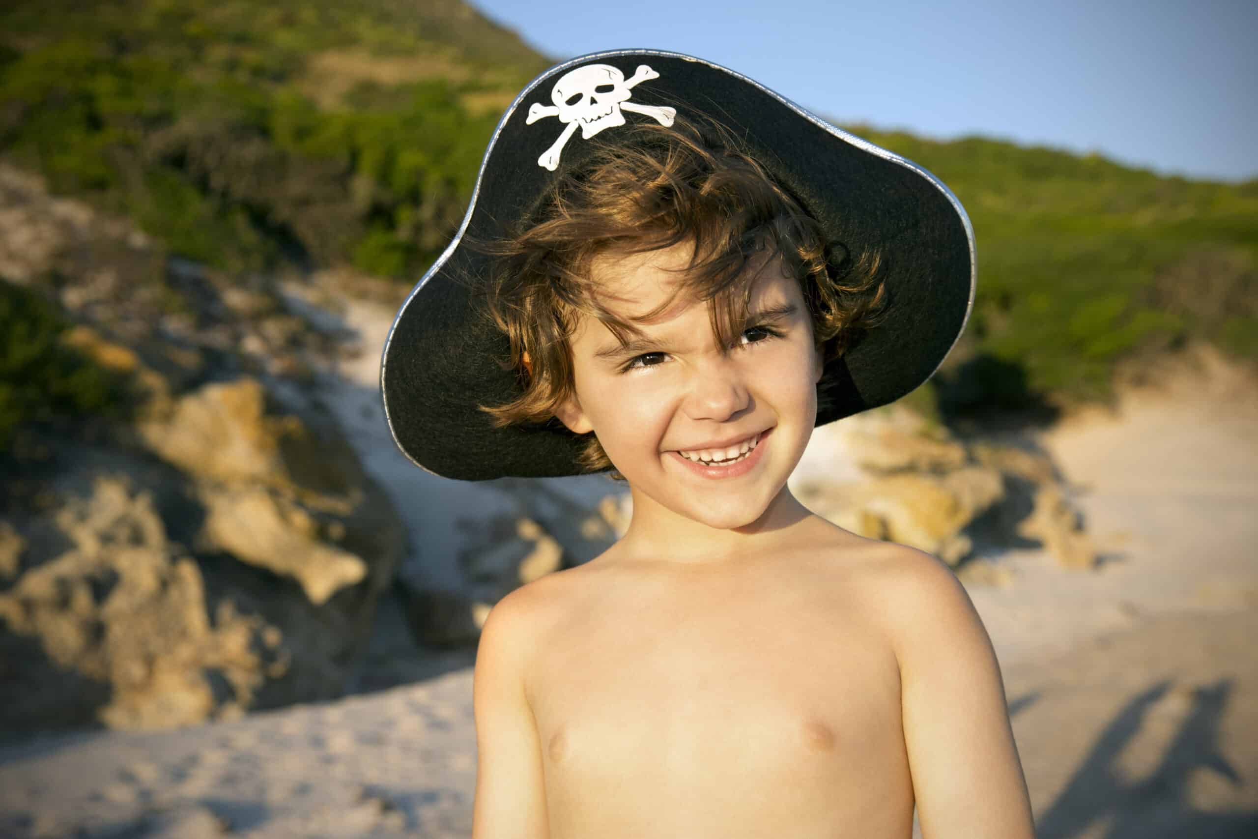 Ein kleines Kind lächelt in die Kamera und trägt dabei einen Piratenhut mit Totenkopf-Motiv. Das Kind ist oben ohne und steht an einem sonnigen Strand mit felsigem Gelände und Grün im Hintergrund. © Fotografie Tomas Rodriguez