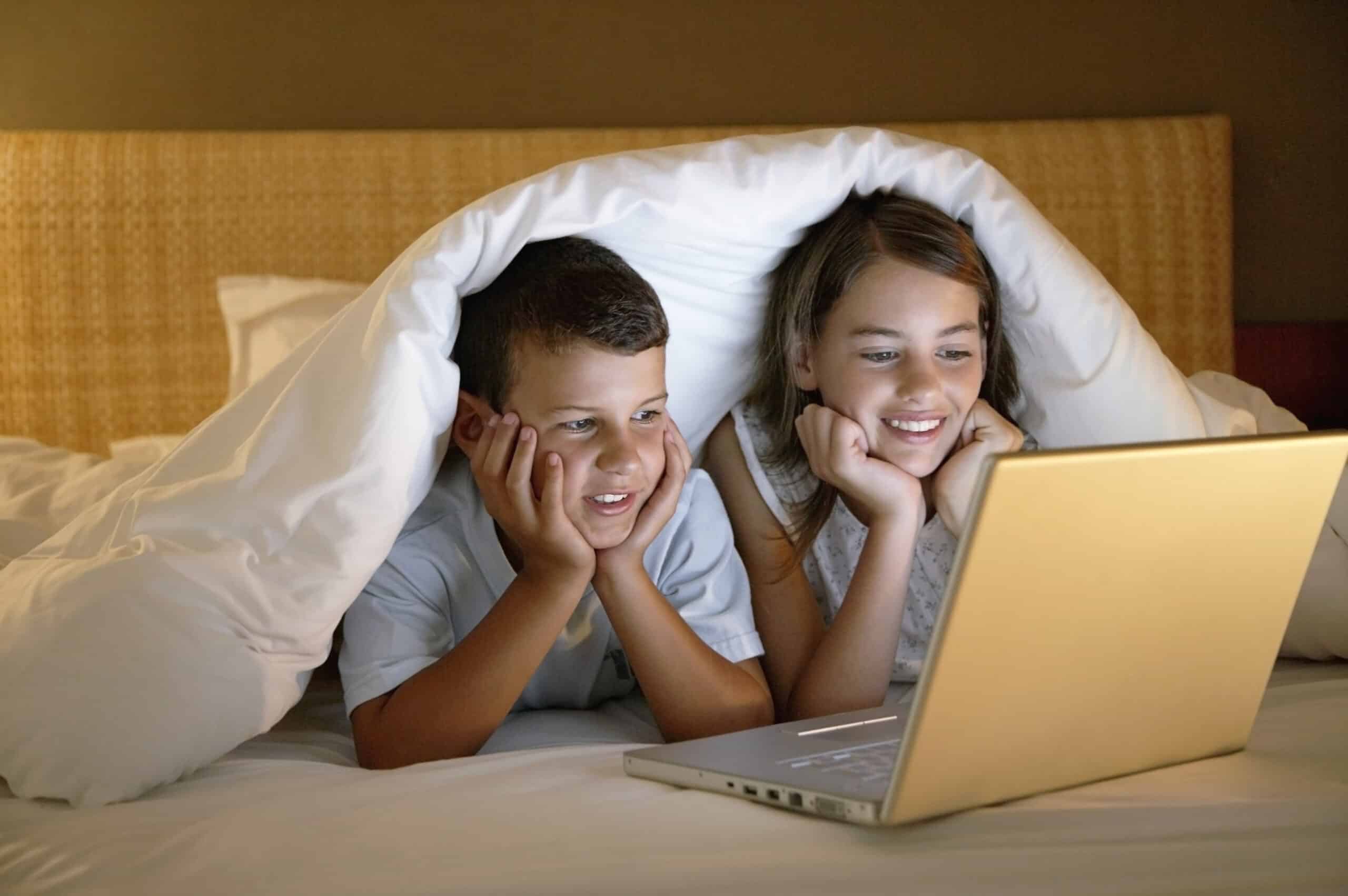 Zwei Kinder liegen auf einem Bett, auf ihre Ellenbogen gestützt, unter einer weißen Decke. Sie schauen auf einen Laptop-Bildschirm vor ihnen, lächeln und wirken interessiert. Der Raum ist schwach beleuchtet, was eine gemütliche Atmosphäre schafft. © Fotografie Tomas Rodriguez