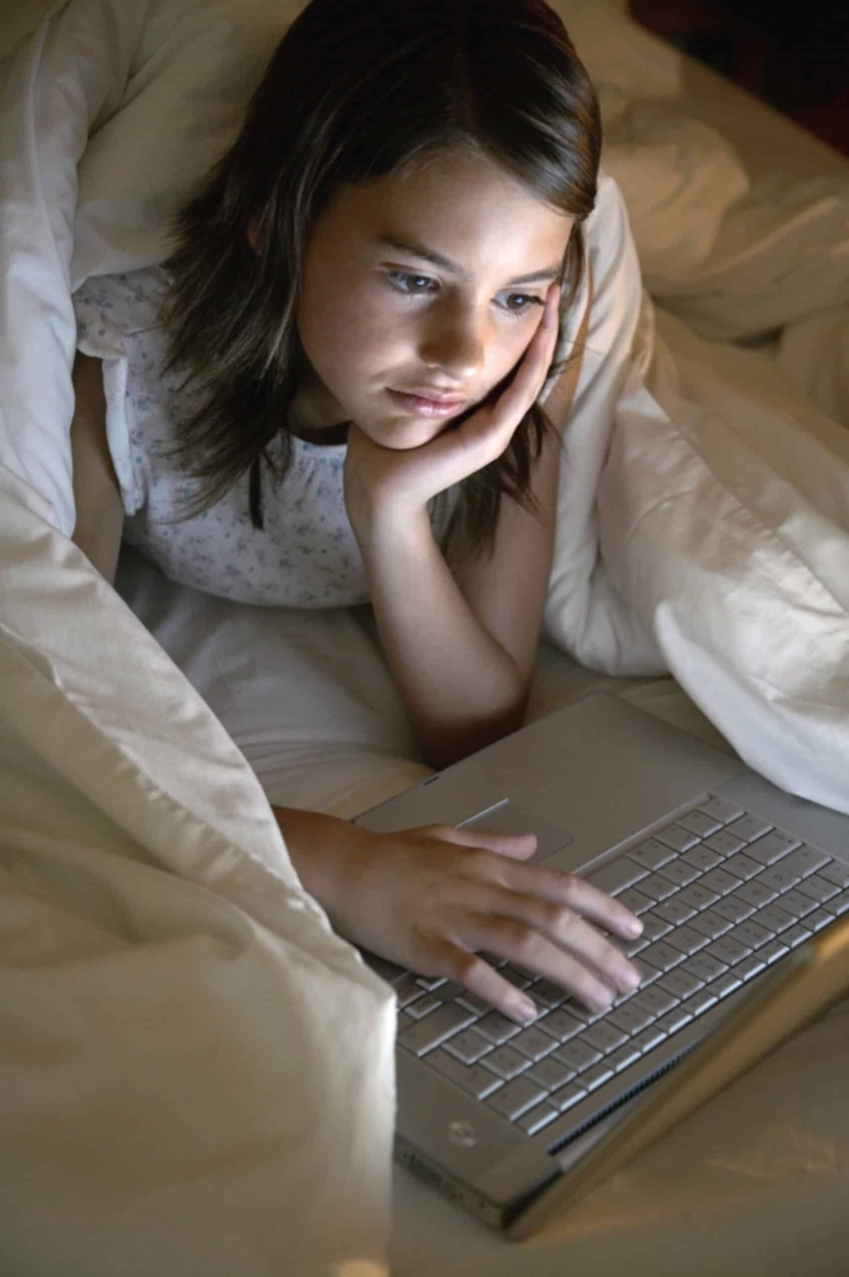 Ein junges Mädchen liegt unter einer weichen weißen Bettdecke im Bett, auf die Ellenbogen gestützt, und schaut auf einen Laptop. Sie stützt ihren Kopf auf eine Hand und tippt mit der anderen. Sie scheint sich intensiv mit dem zu beschäftigen, was auf dem Bildschirm angezeigt wird. © Fotografie Tomas Rodriguez