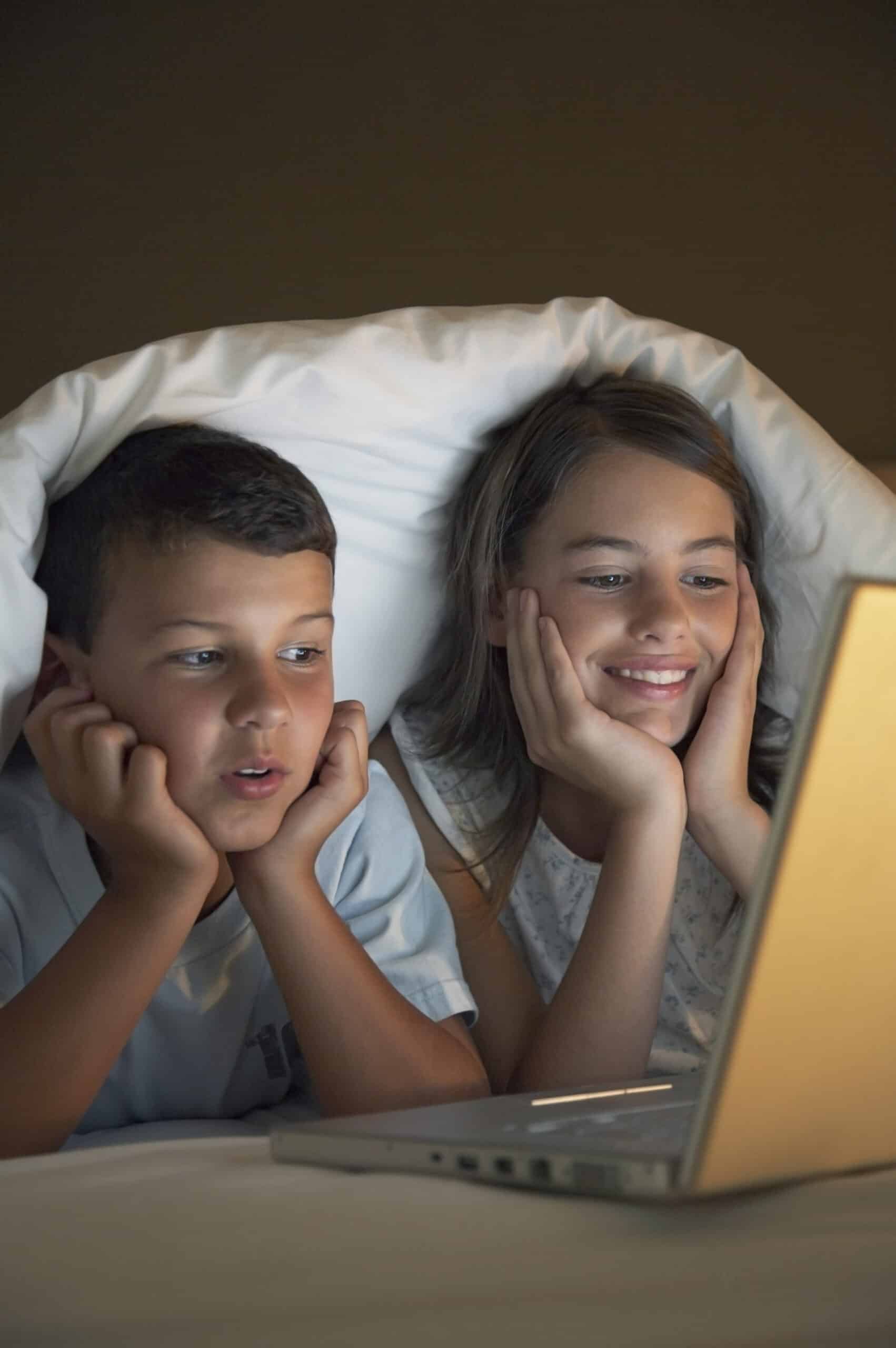 Zwei Kinder, ein Junge und ein Mädchen, liegen unter einer Decke und schauen gespannt auf den Bildschirm eines Laptops. Das warme Licht des Bildschirms erhellt ihre Gesichter und beide haben einen Ausdruck der Neugier und Freude. © Fotografie Tomas Rodriguez