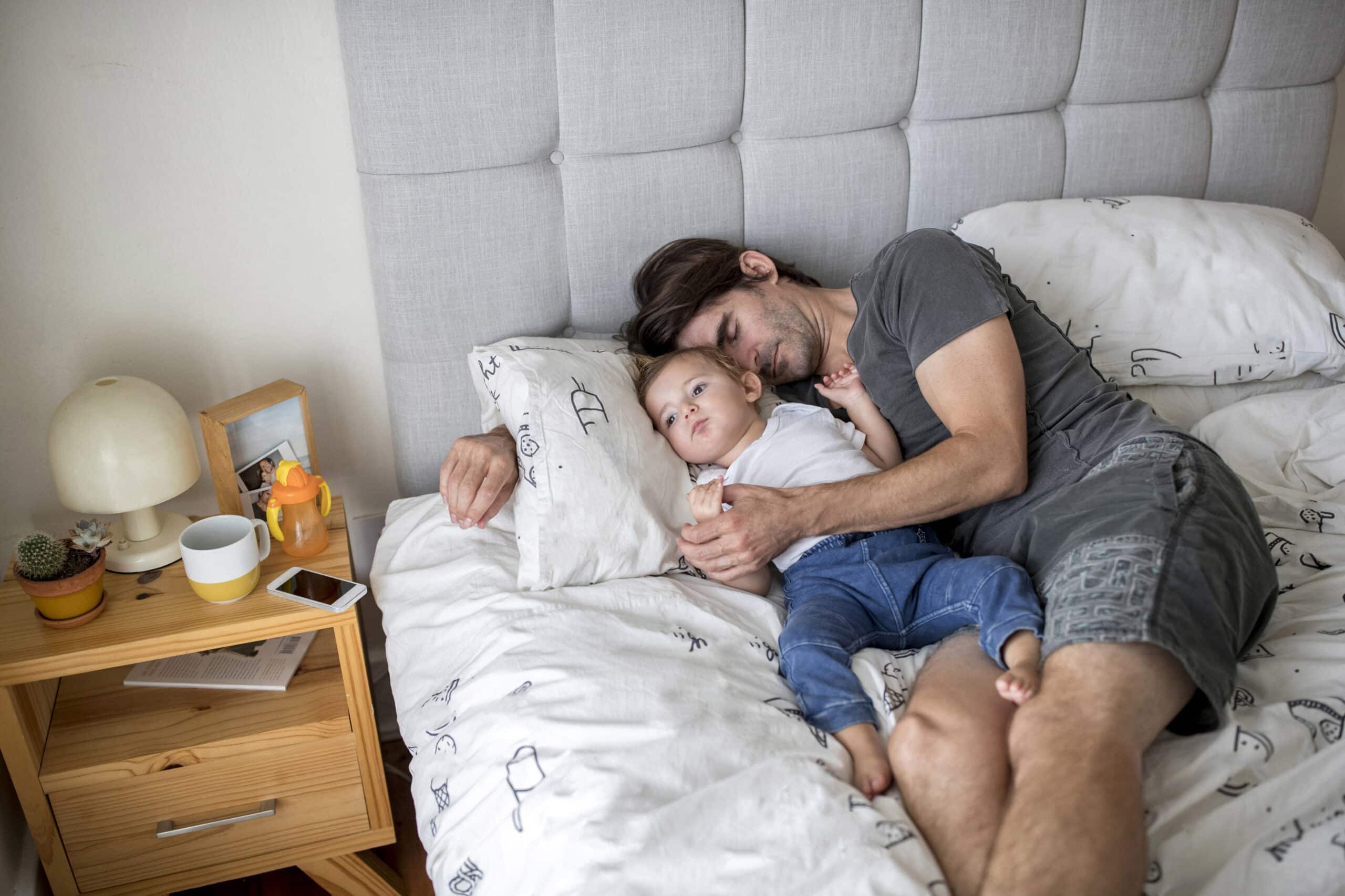 Ein Mann mit dunklem Haar und Bart, der ein graues T-Shirt trägt, liegt auf einem Bett und kuschelt ein kleines Kind, das ein weißes Hemd und eine blaue Hose trägt. Sie ruhen beide auf weißen Kissen mit gemusterter Bettwäsche. Auf einem hölzernen Nachttisch liegen verschiedene Gegenstände, darunter eine Lampe und ein Telefon. © Fotografie Tomas Rodriguez