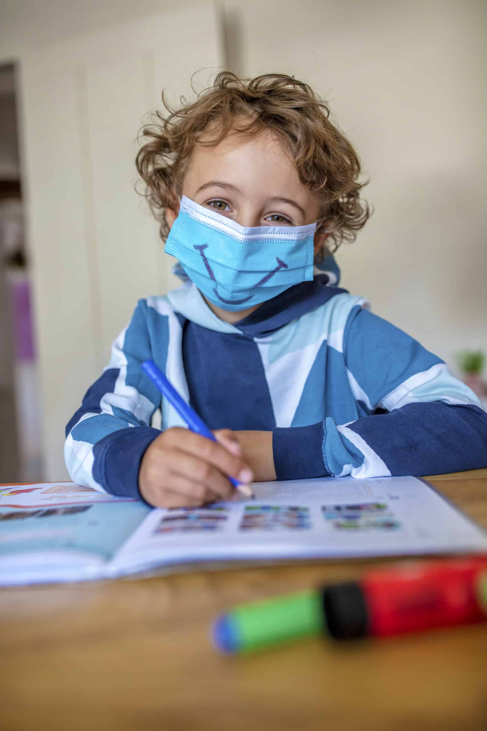Ein kleines Kind mit lockigem Haar und einer Gesichtsmaske mit einem lächelnden Gesicht sitzt an einem Tisch und schreibt mit einem blauen Stift in ein Arbeitsbuch. Das Kind trägt ein gestreiftes blaues Sweatshirt. Im Vordergrund liegen bunte Marker auf dem Tisch. © Fotografie Tomas Rodriguez