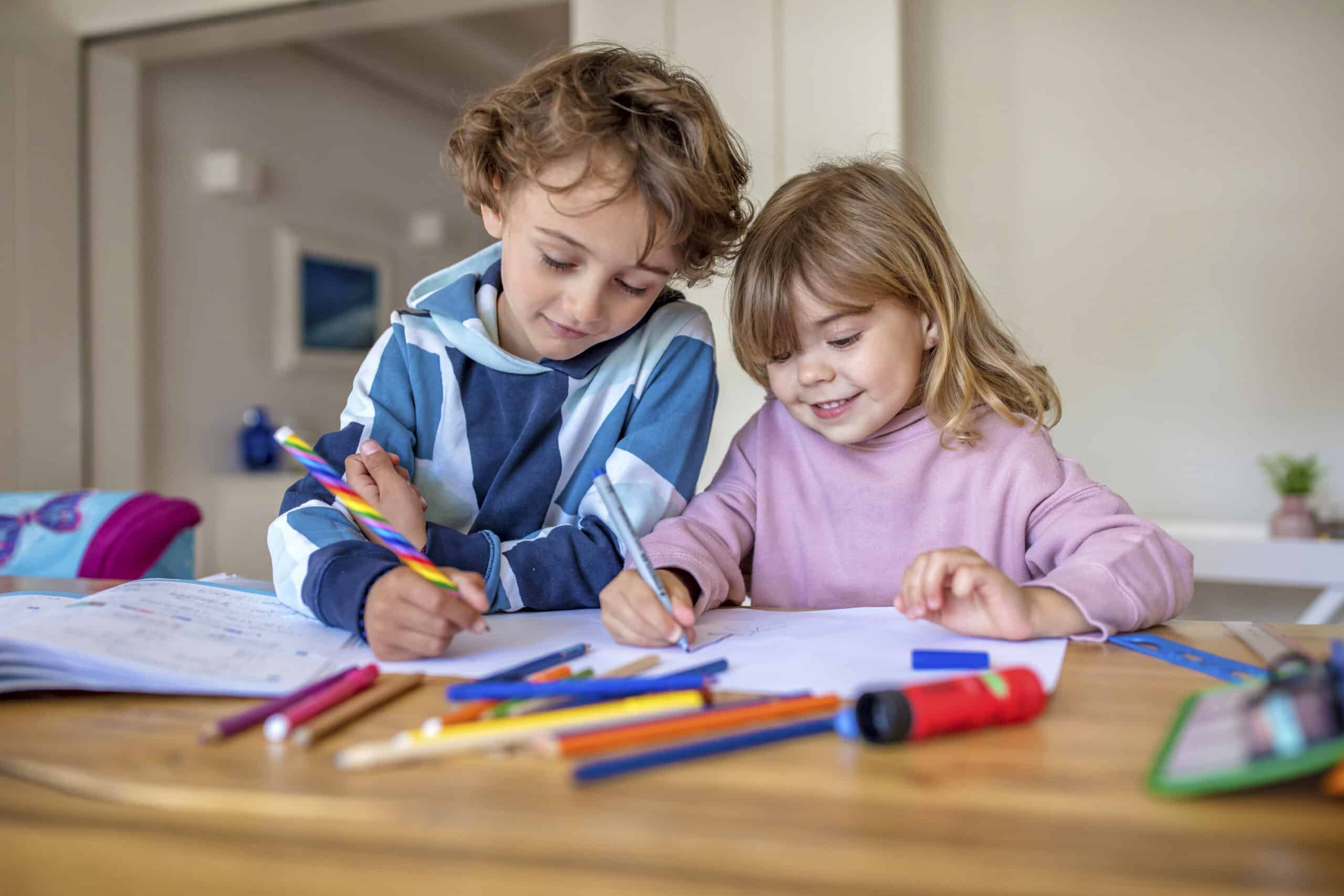 Zwei kleine Kinder, eines mit lockigem Haar und einem blau gestreiften Kapuzenpullover, das andere mit glattem Haar und einem rosa Pullover, sitzen an einem Tisch und malen mit Buntstiften und Markern auf weißem Papier. Verschiedene Malutensilien liegen verstreut auf dem Tisch. © Fotografie Tomas Rodriguez
