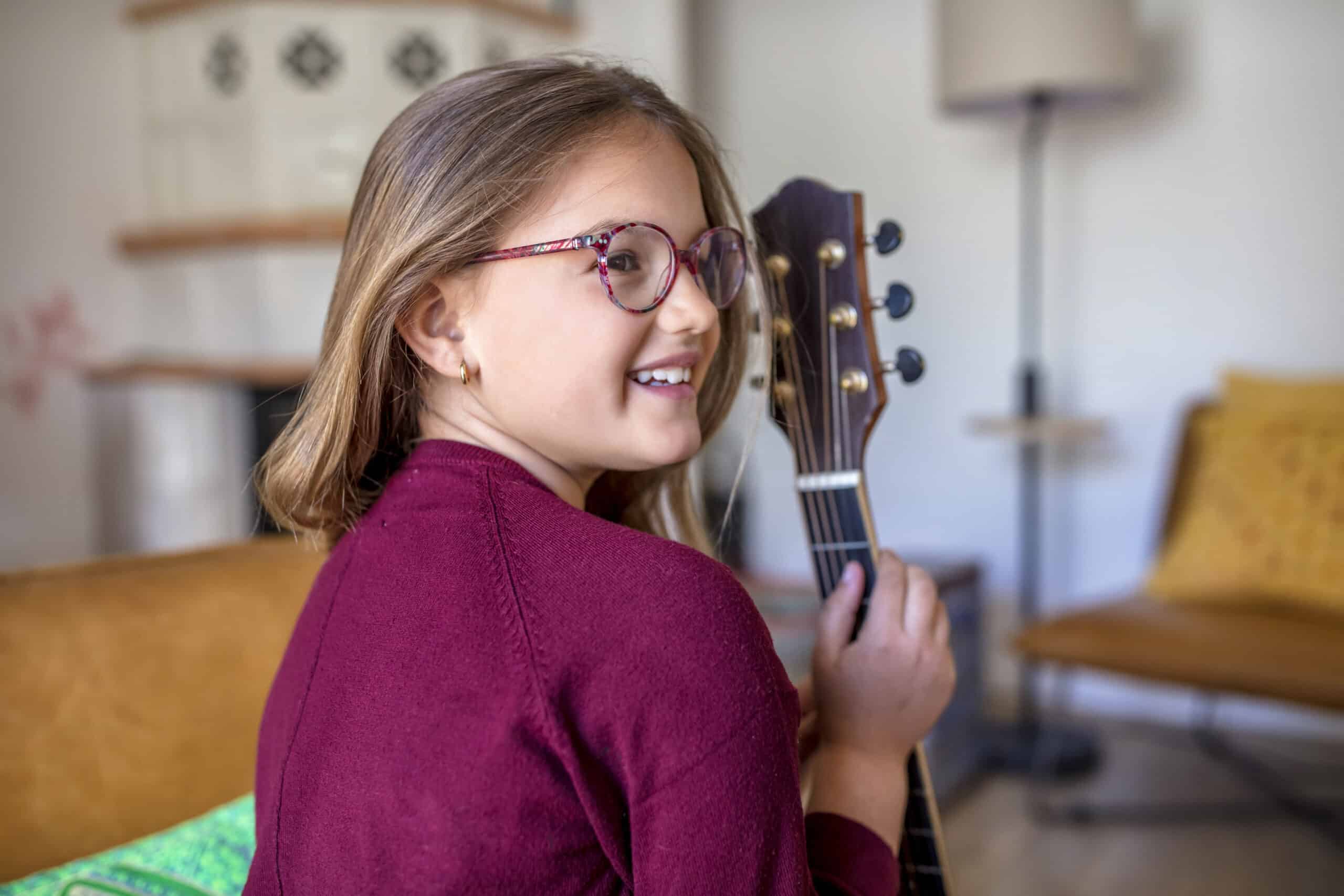 Ein junges Mädchen mit Brille und einem kastanienbraunen Pullover sitzt auf einem Sofa und hält eine Akustikgitarre in der Hand. Sie lächelt und blickt über ihre Schulter zurück. Im Hintergrund ist ein gemütliches Wohnzimmer mit einer Lampe, einem gelben Kissen und Holzmöbeln zu sehen. © Fotografie Tomas Rodriguez