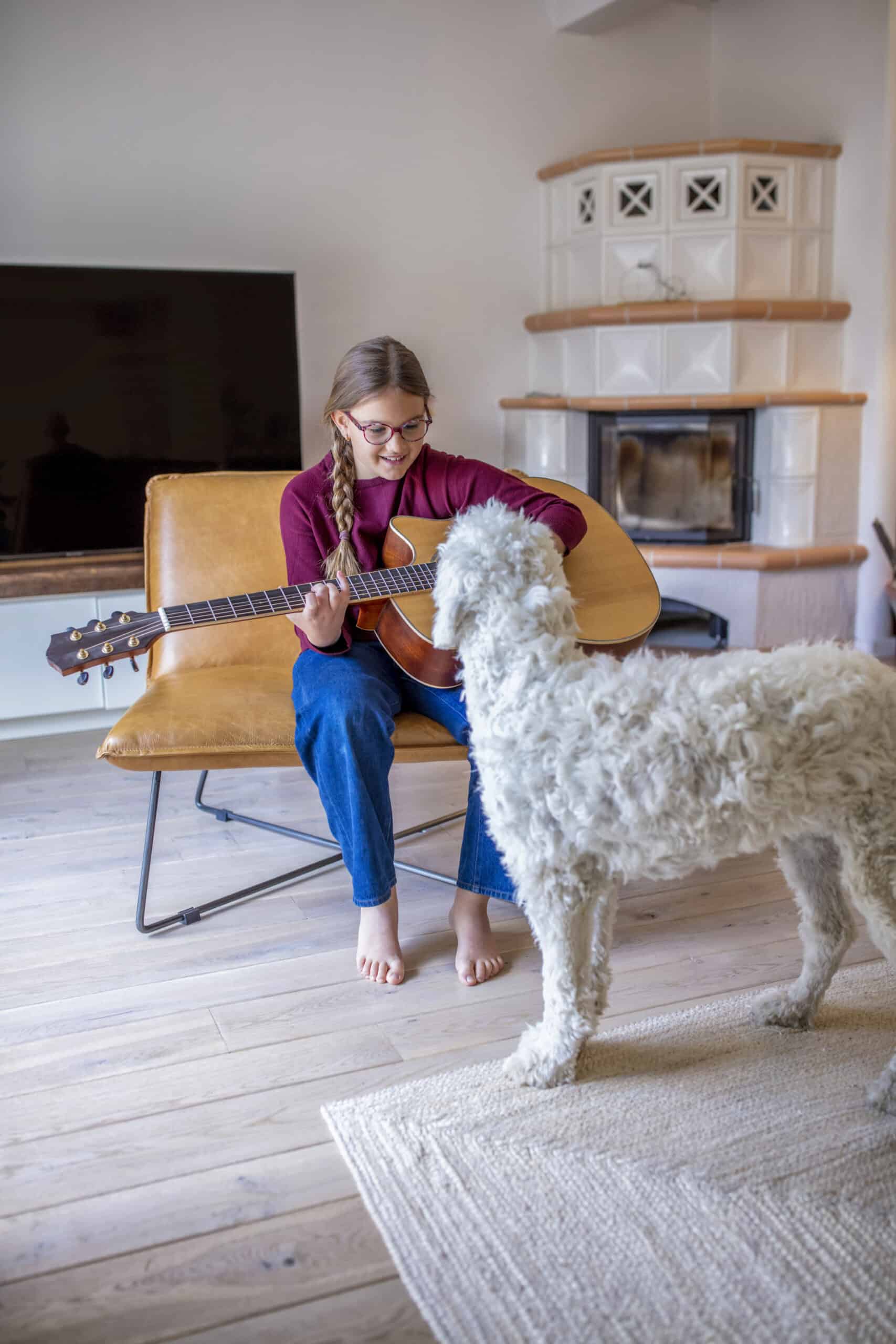 Eine Person mit geflochtenen Haaren, die ein rotes Langarmshirt und blaue Jeans trägt, sitzt auf einem braunen Sofa und spielt Akustikgitarre. Ein Hund mit lockigem Fell steht in der Nähe auf einem Teppich und blickt in die Richtung der Person. Im Hintergrund sind ein Fernseher und ein Kamin zu sehen. © Fotografie Tomas Rodriguez