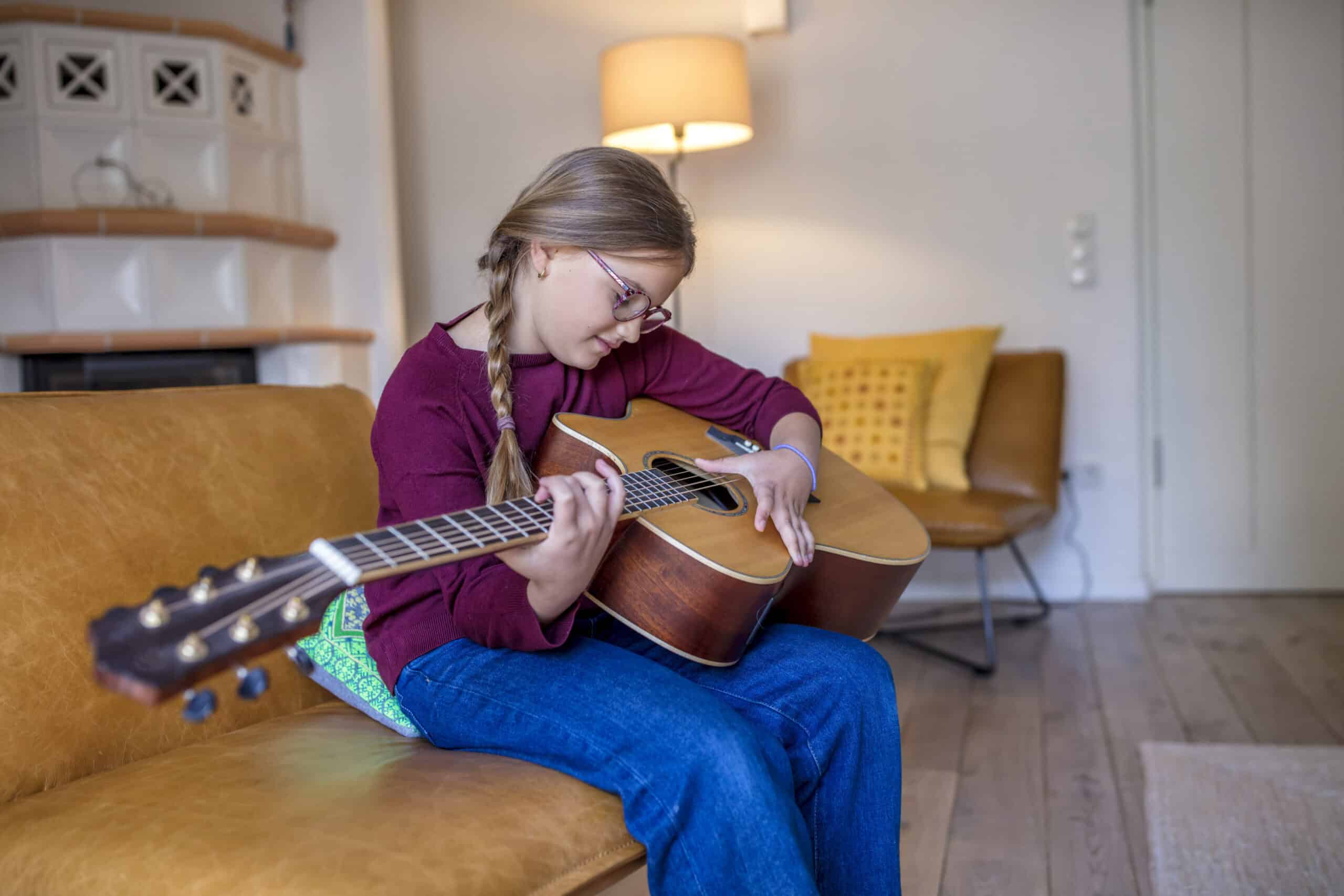 Ein junges Mädchen mit Brille und langen Zöpfen spielt Akustikgitarre, während es auf einem Sofa in einem gemütlichen Wohnzimmer sitzt. Im Hintergrund sind eine Lampe und ein Stuhl mit Kissen zu sehen. Der Raum hat Holzboden und sanfte, warme Beleuchtung. © Fotografie Tomas Rodriguez