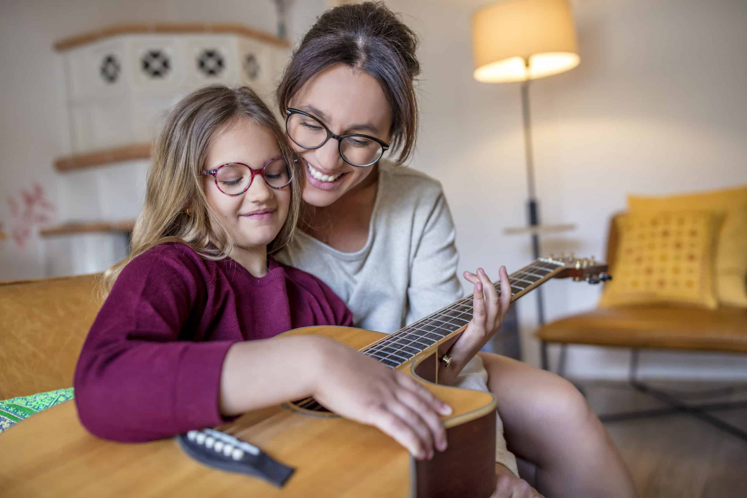 Eine Frau mit Brille und langen Haaren lächelt, während sie einem jungen Mädchen, das ebenfalls eine Brille trägt, beim Spielen einer Akustikgitarre hilft. Sie sitzen zusammen auf einem Sofa in einem gemütlichen Wohnzimmer mit warmer Beleuchtung und einer Lampe im Hintergrund. © Fotografie Tomas Rodriguez